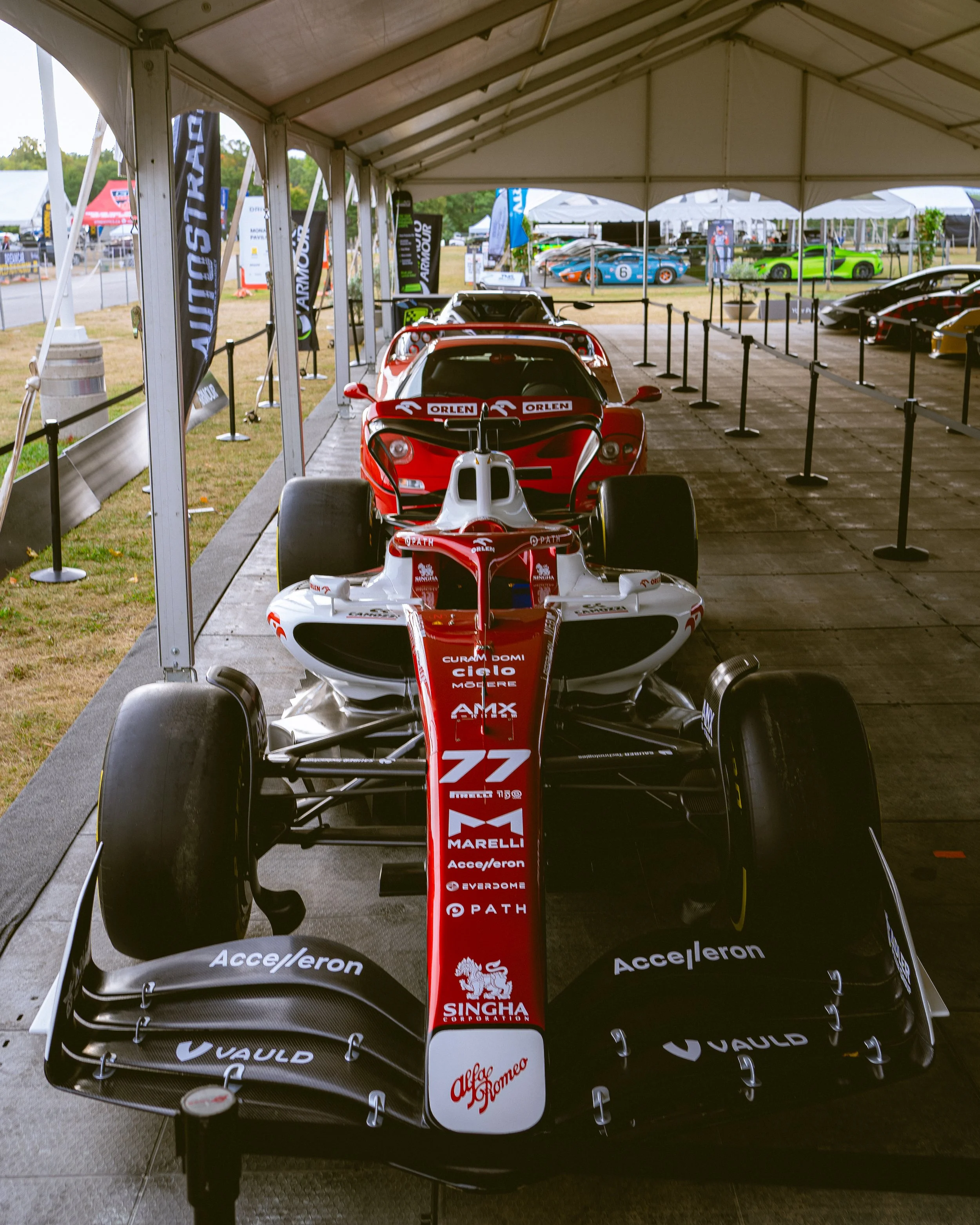 Multiple race cars displayed under a large canopy at a motorsport event, with several other cars and tents visible in the background.