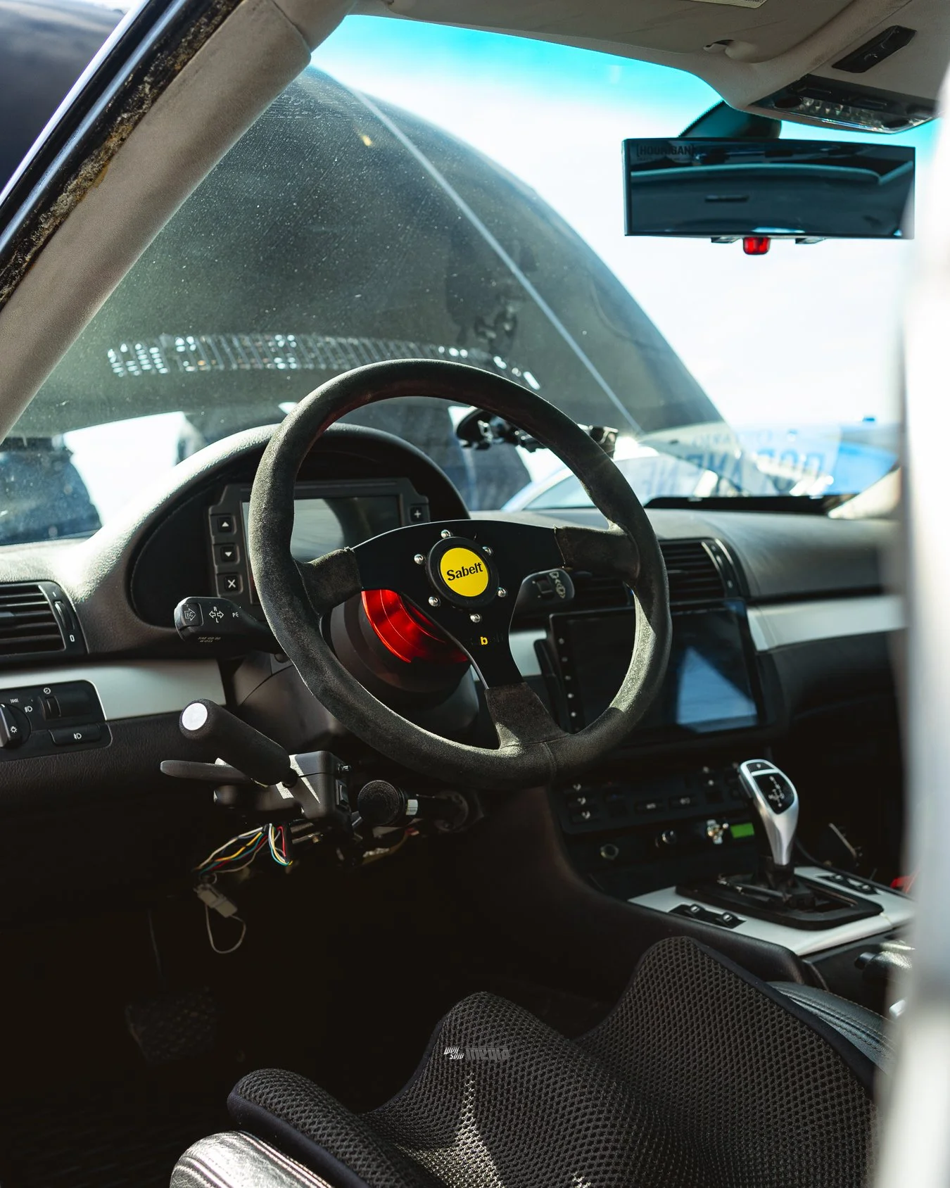 Interior of a vehicle, showing a racing steering wheel with a yellow center labeled 'Sabelt', dashboard controls, and a digital display. The windshield is dusty and a monitor is mounted on the ceiling.