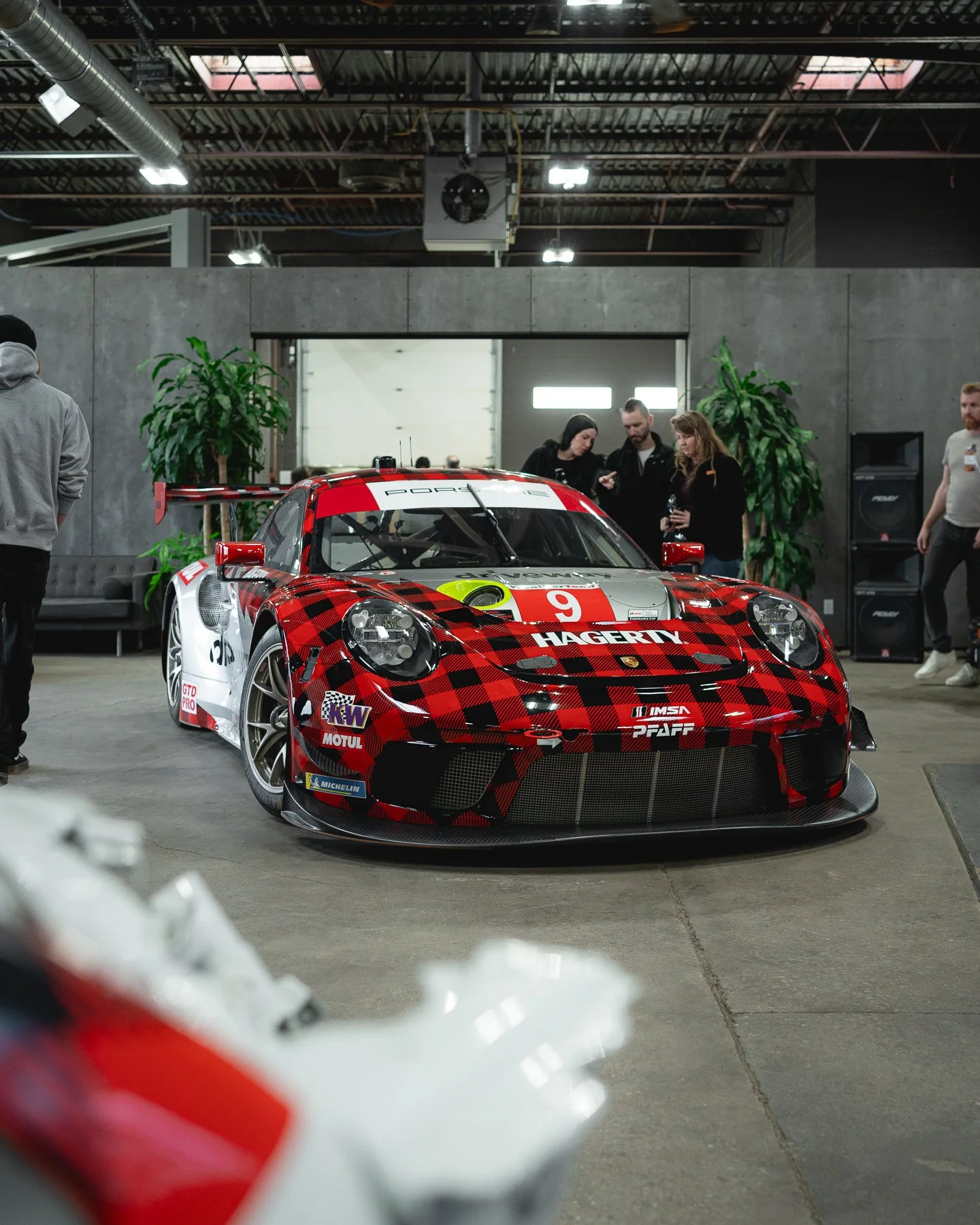 A red and black race car with sponsorship decals displayed indoors, surrounded by people and potted plants, in an industrial-style setting.