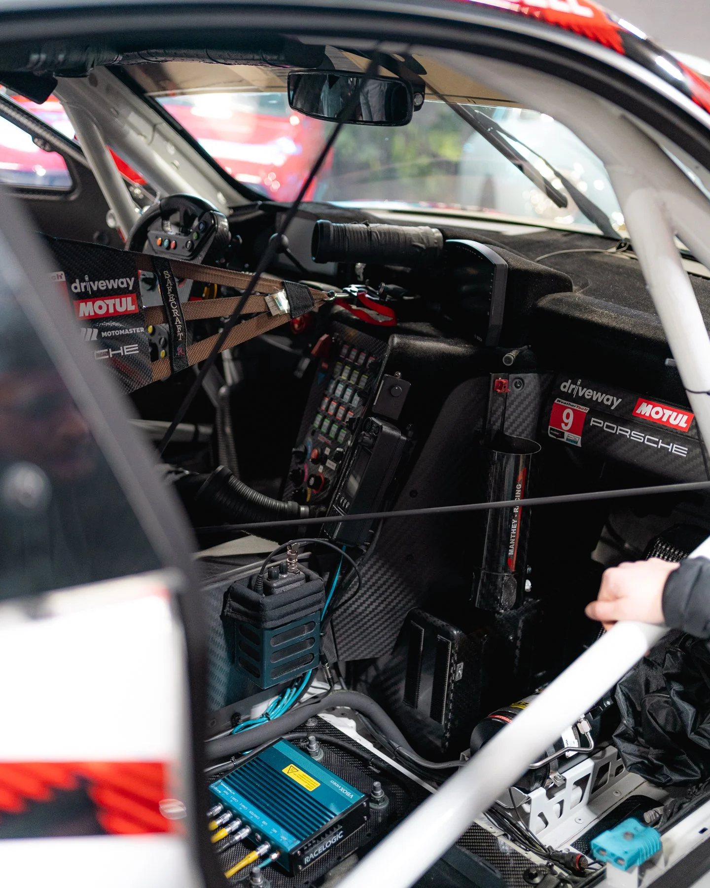 Interior of a race car cockpit showing steering wheel, dashboard with multiple buttons, racing harness, communication device, and various racing equipment and branding.
