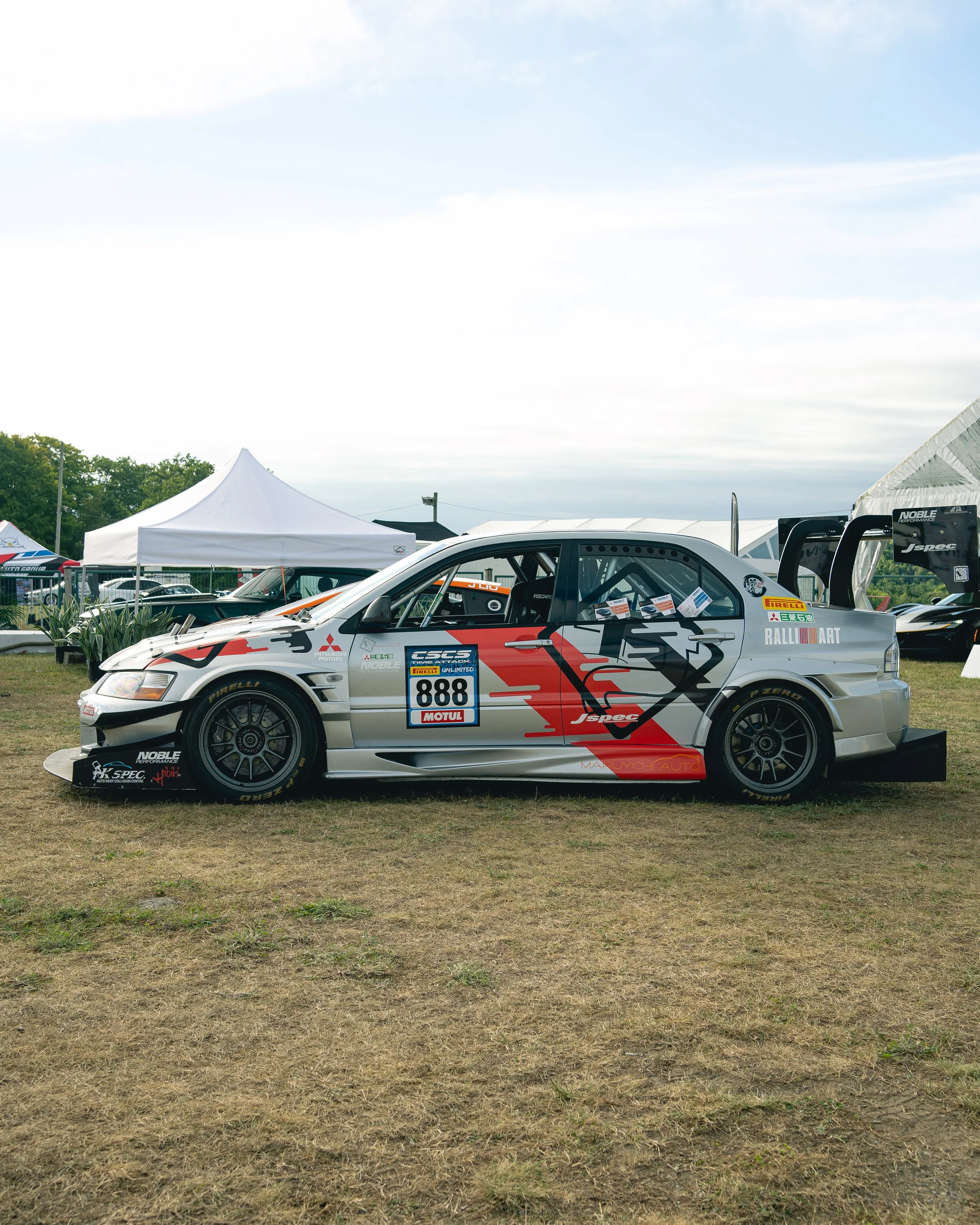 Silver race car with red and black graphics and the number 888, parked on grass at an outdoor event with white tents and other racing cars in the background.