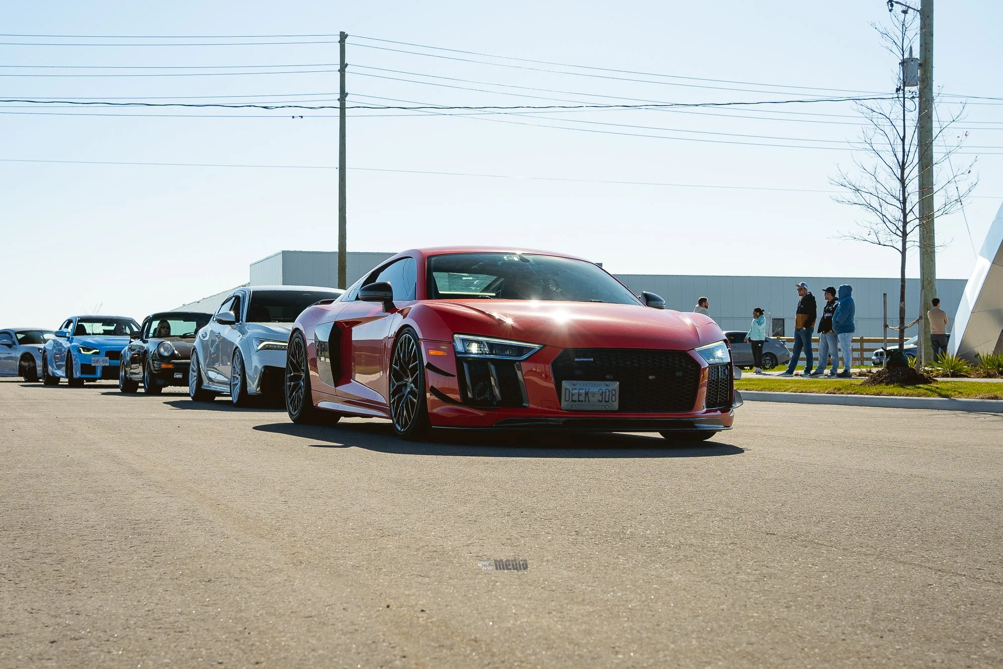 Red sports car parked in an outdoor parking lot with other vehicles and people walking in the background. Clear sky and utility poles visible overhead.