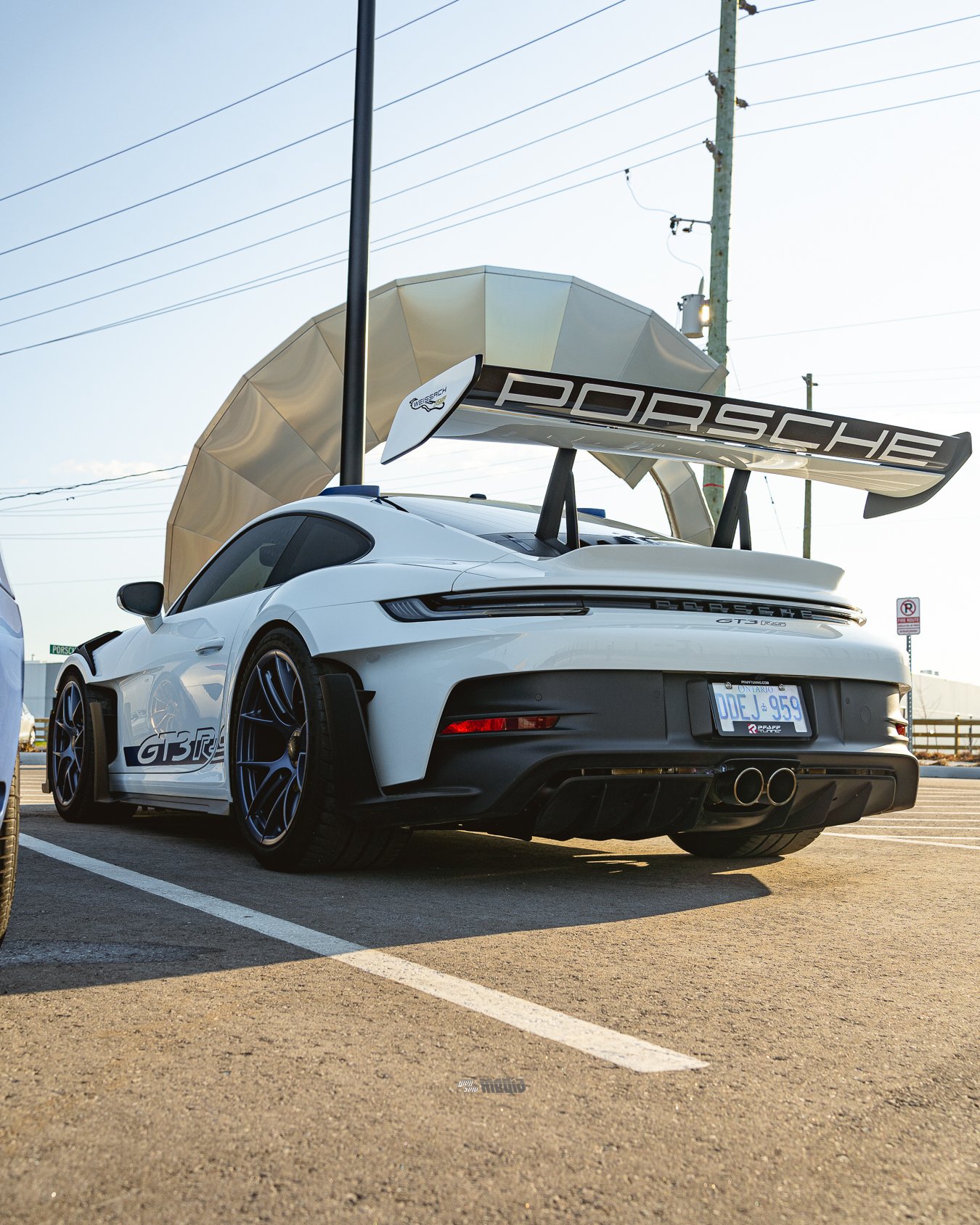 A white Porsche 911 GT3 RS sports car parked in a parking lot during daytime. The car features a large rear wing with the Porsche logo, black wheels, and dual exhaust pipes. There are power lines and utility poles in the background, and the sky is cl