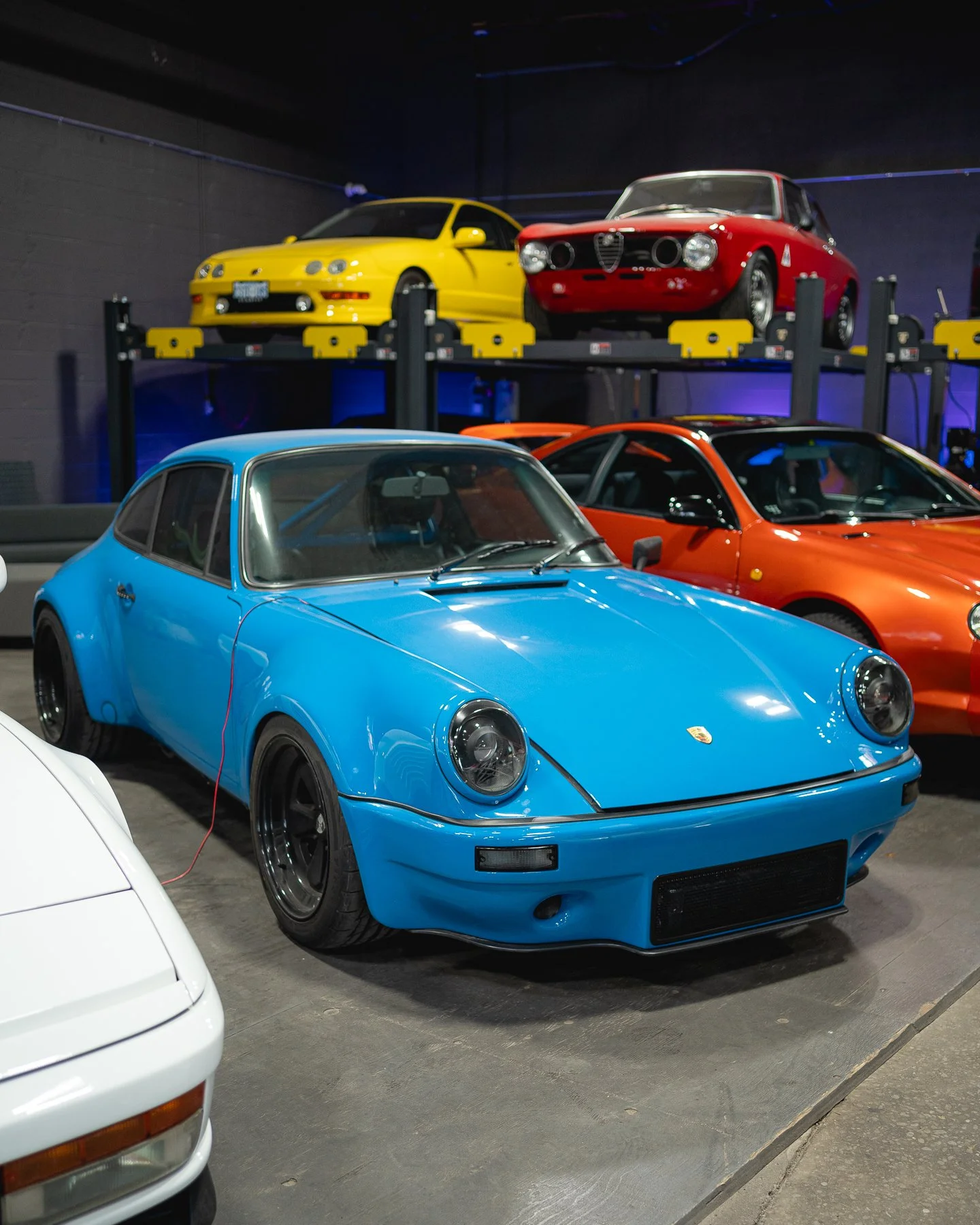 A blue vintage Porsche parked indoors among other cars, with additional cars displayed on a raised platform in the background.