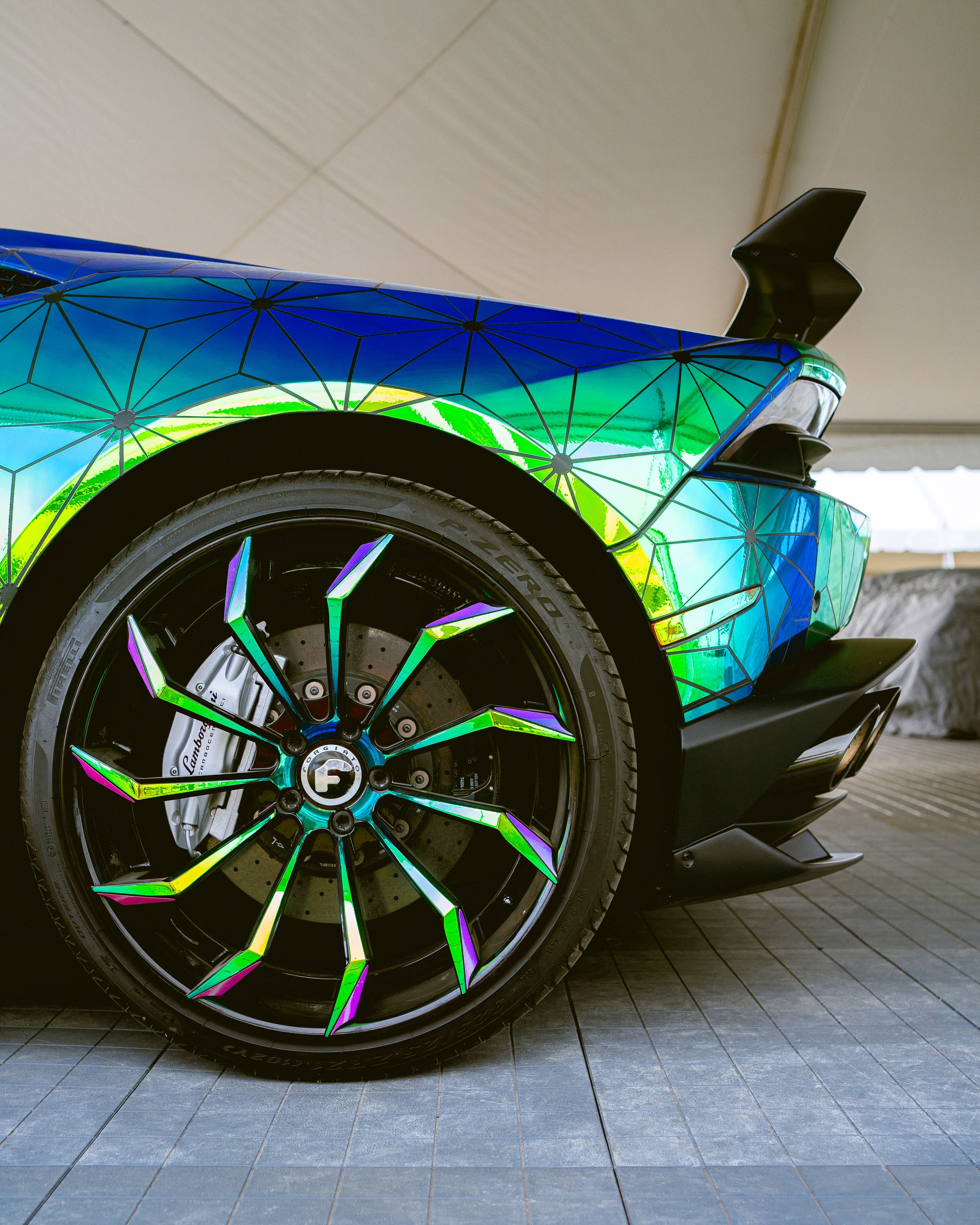 Close-up of a sports car with a multicolored geometric design on the body, showcasing the front wheel, brake caliper, and rear wing, parked on a tiled surface.