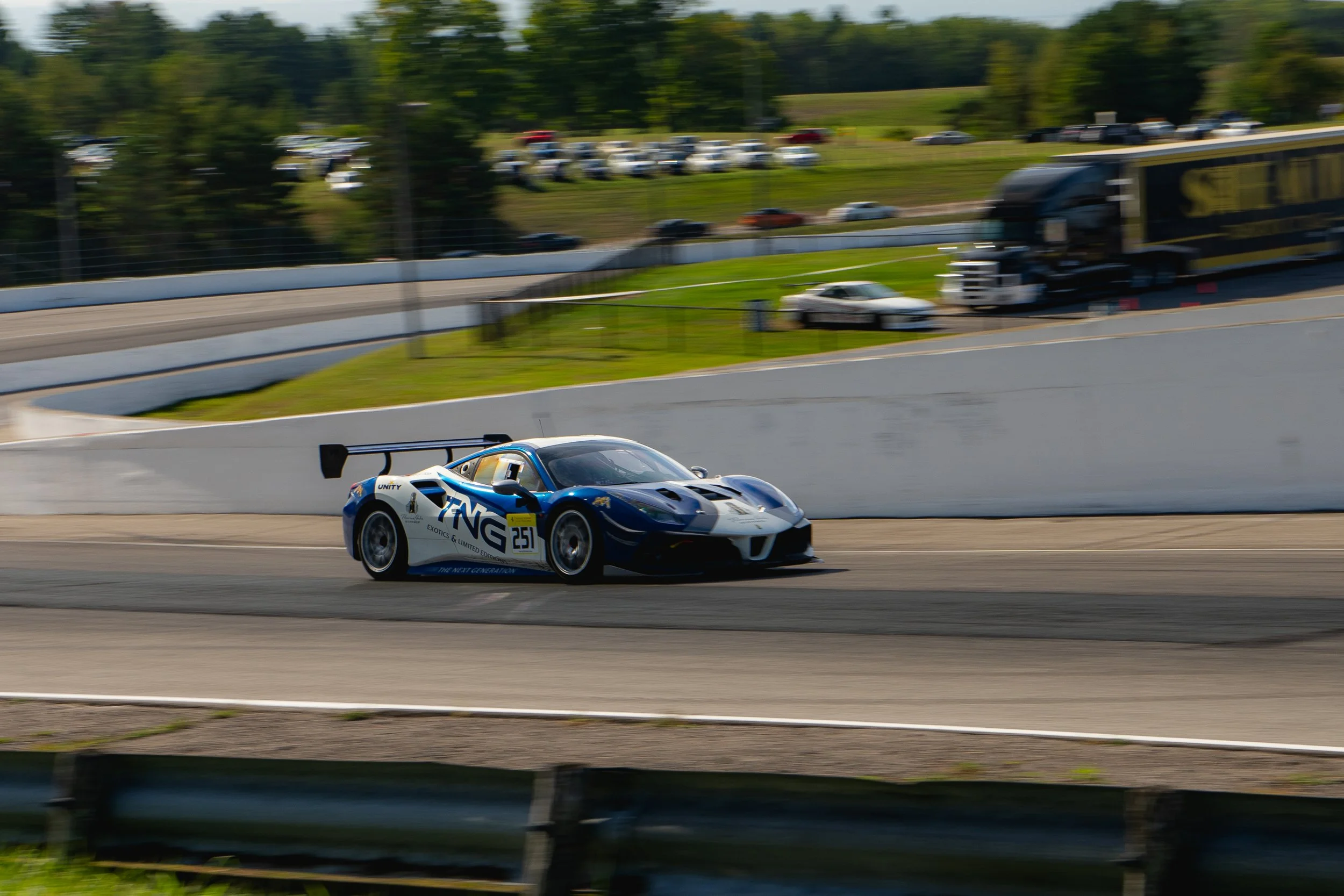 Blue and white race car speeding on a race track, with support vehicles and trees in the background.