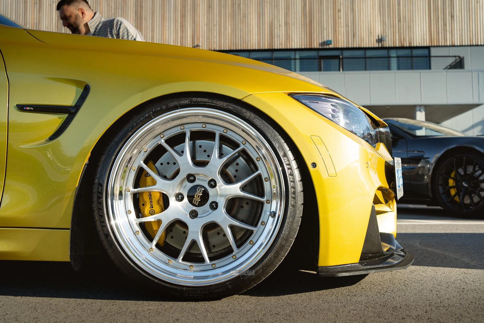 Close-up of a yellow sports car's front wheel with polished silver rim and brake caliper, parked outdoors with a man looking down at it and a modern building in the background.