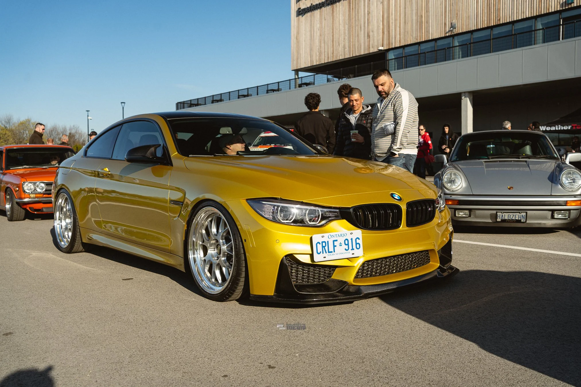 A yellow BMW sports car on display at a car show, with a group of people gathered around, looking at it and talking. The background includes a modern building and several other cars.