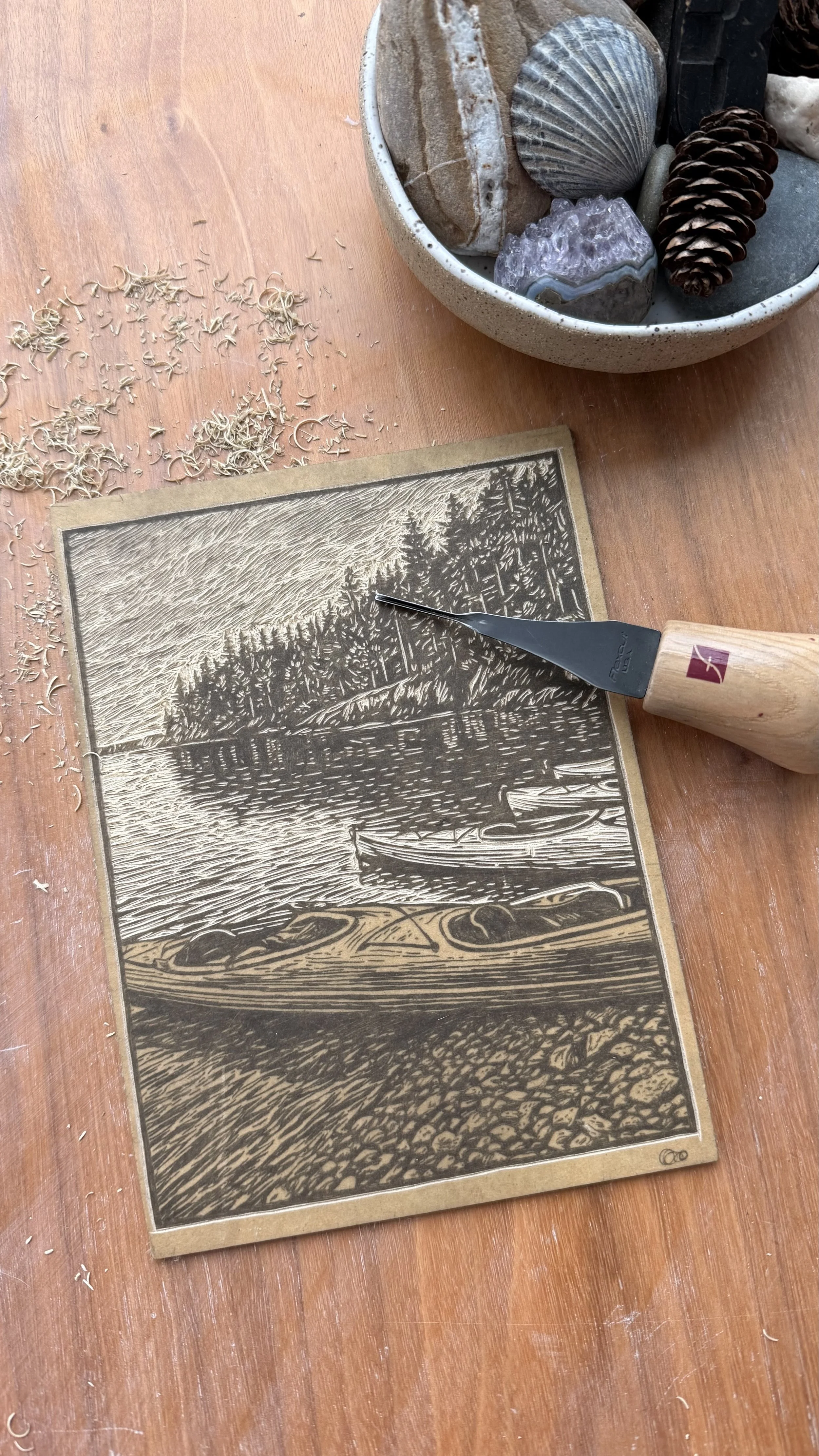 Linocut print of kayaks resting along a rocky shoreline with forest across the water, hand printed from a carved linoleum block in the On The Water collection.