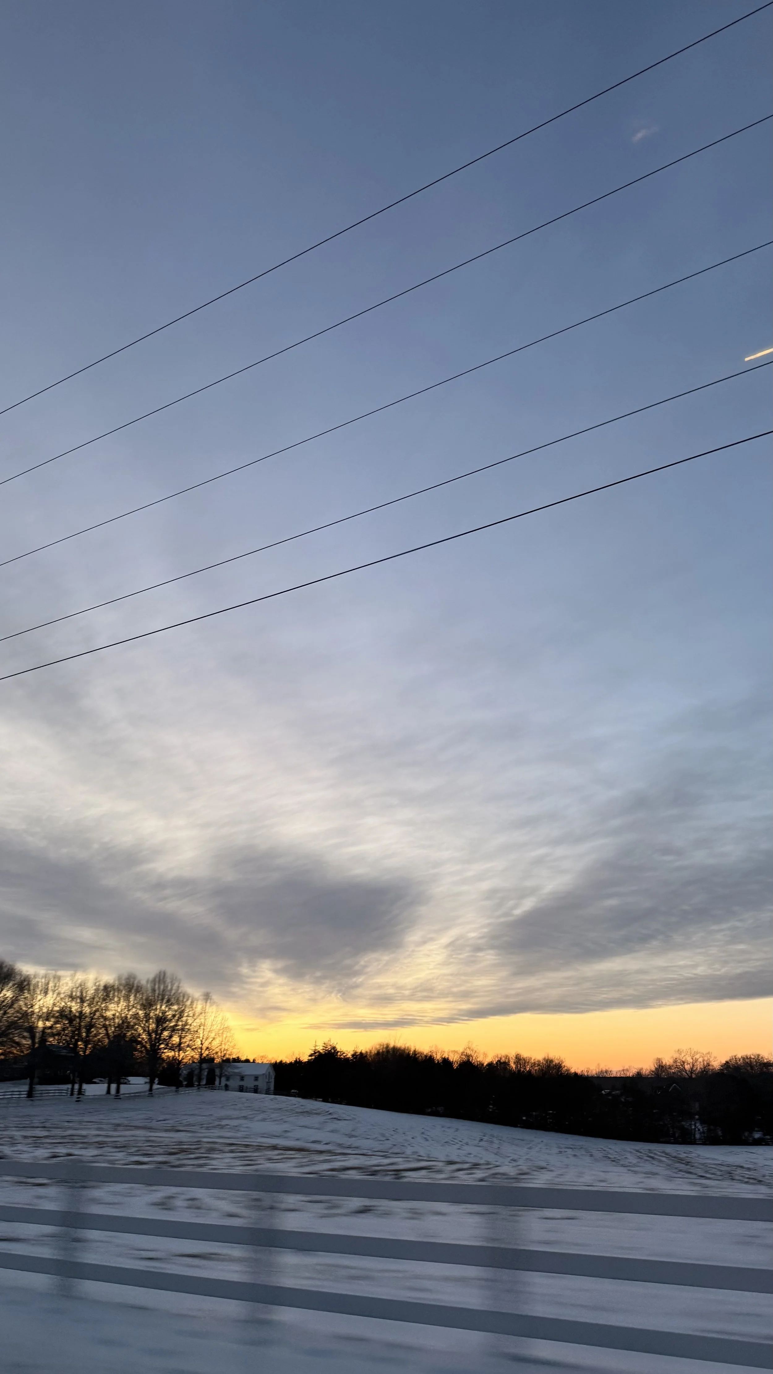 Winter sunset over a snow-covered field with bare trees on the horizon and power lines stretching across a blue and gold sky.