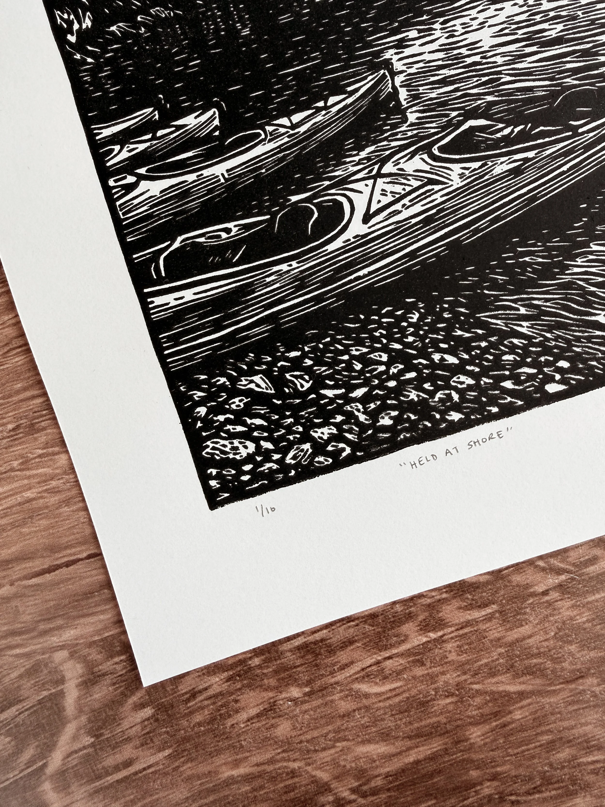Close-up of linocut print showing kayaks at shoreline with rocky foreground, titled Held at Shore