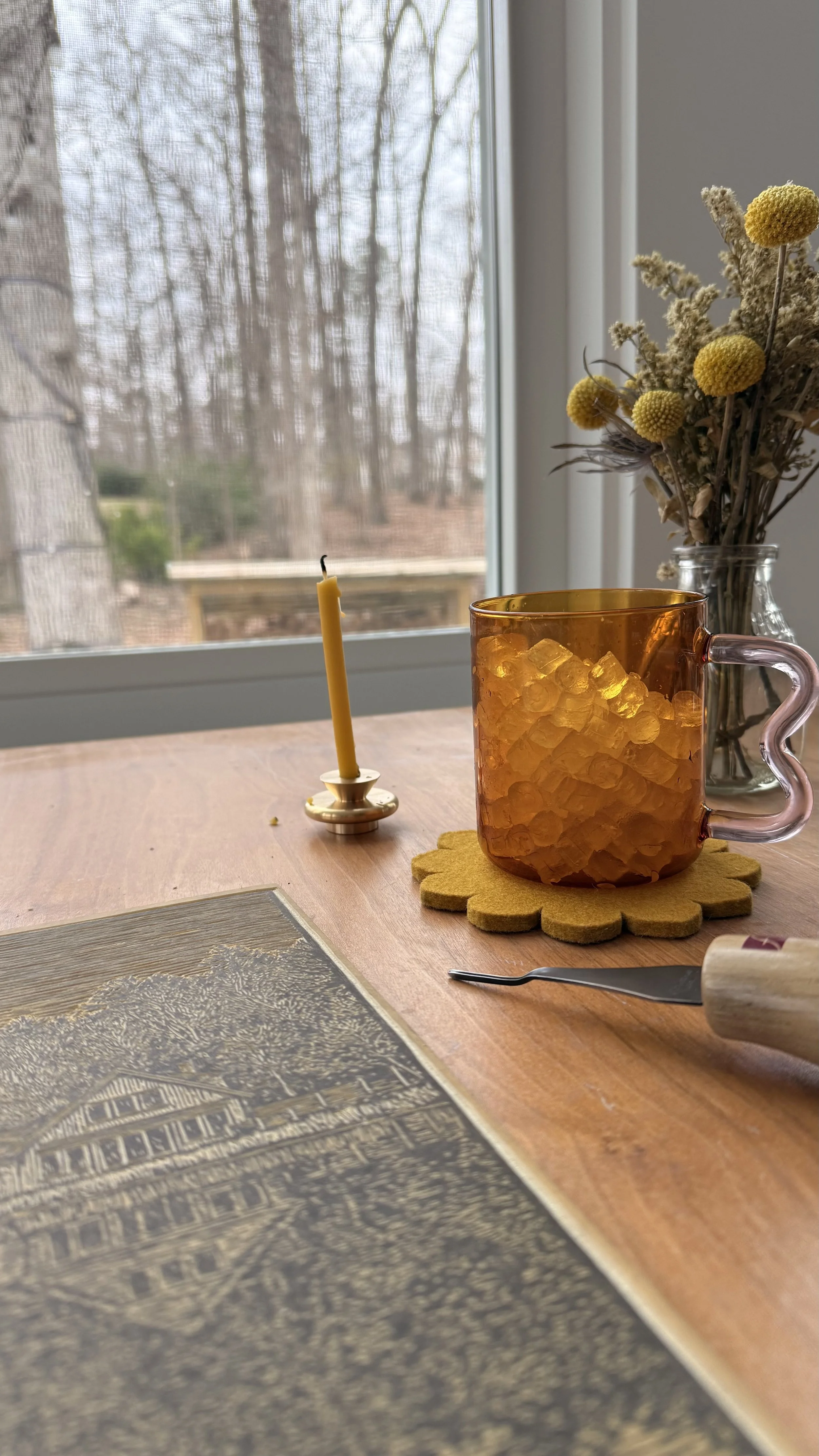 A wooden table by a window holding a linocut block, carving tool, amber glass mug, candle, and a small vase of dried flowers, with winter trees visible outside.