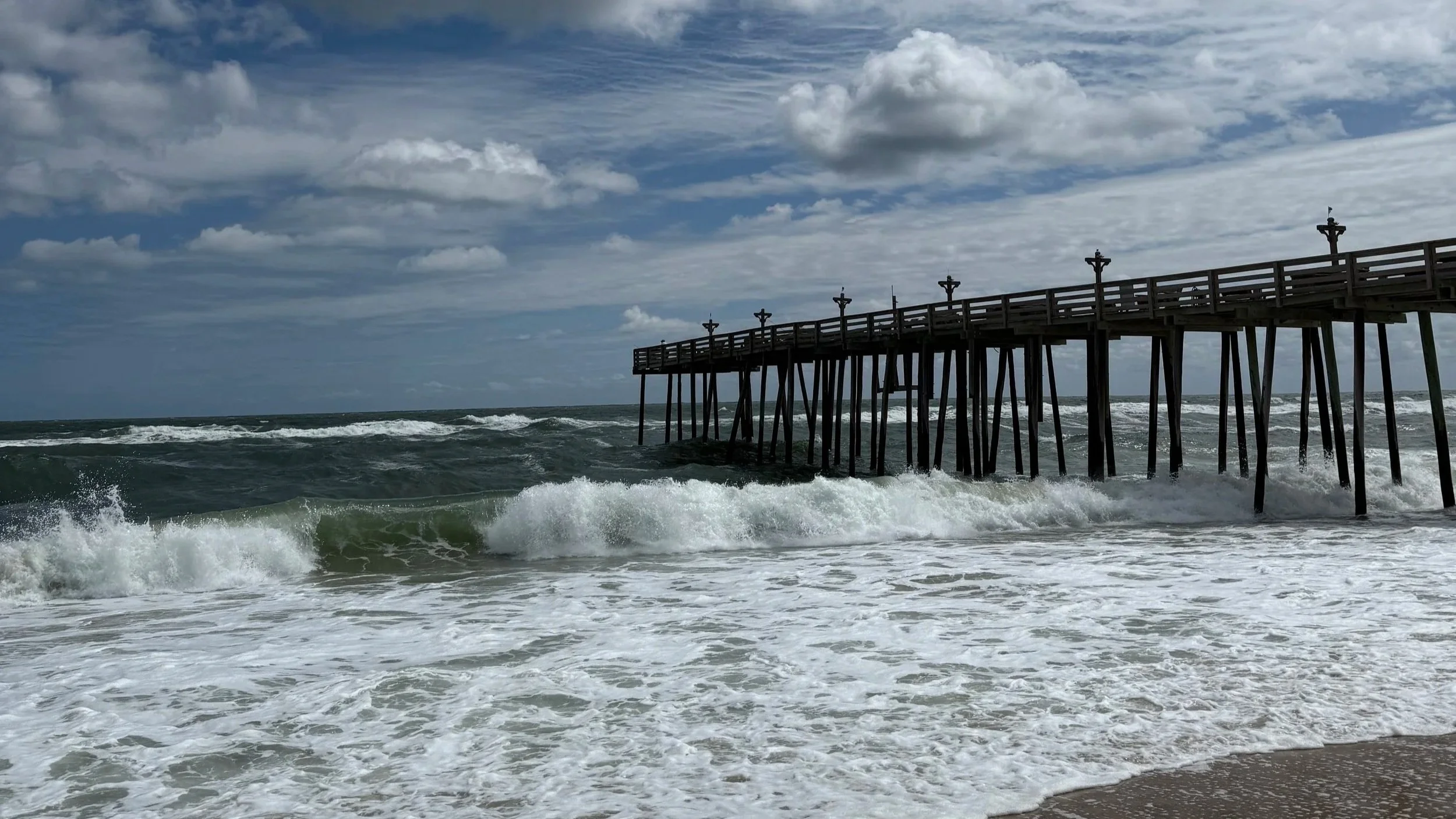 Ocean waves crashing near a wooden pier under a wide sky during a quiet morning in Outer Banks, North Carolina