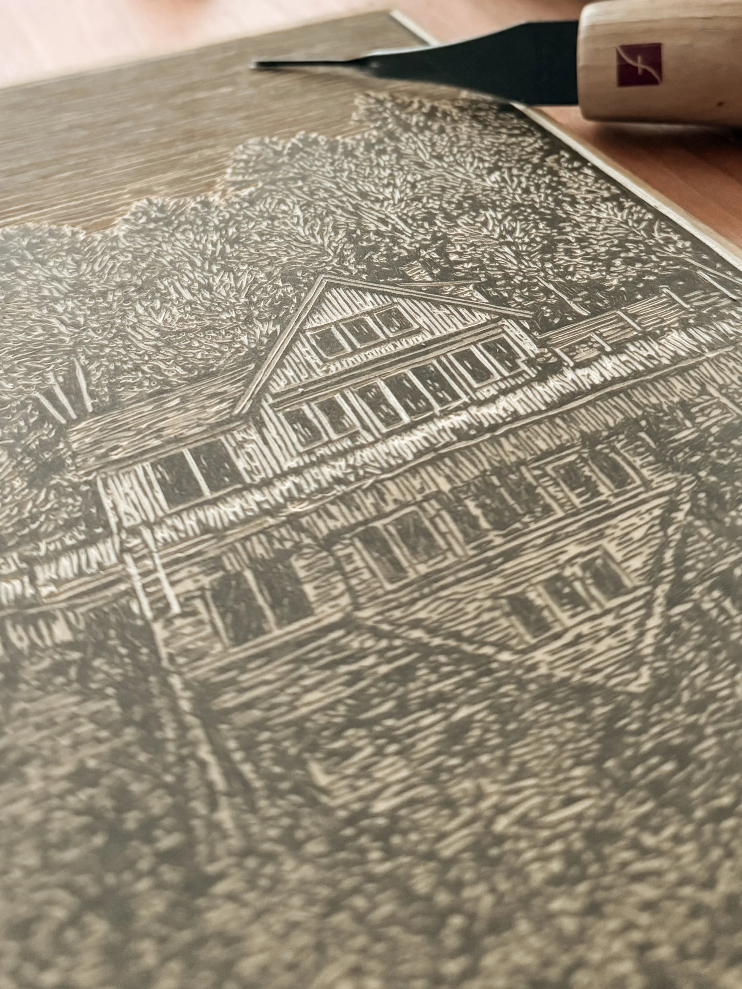 Carving tool resting on linoleum block with detailed lakeside cabin design in progress