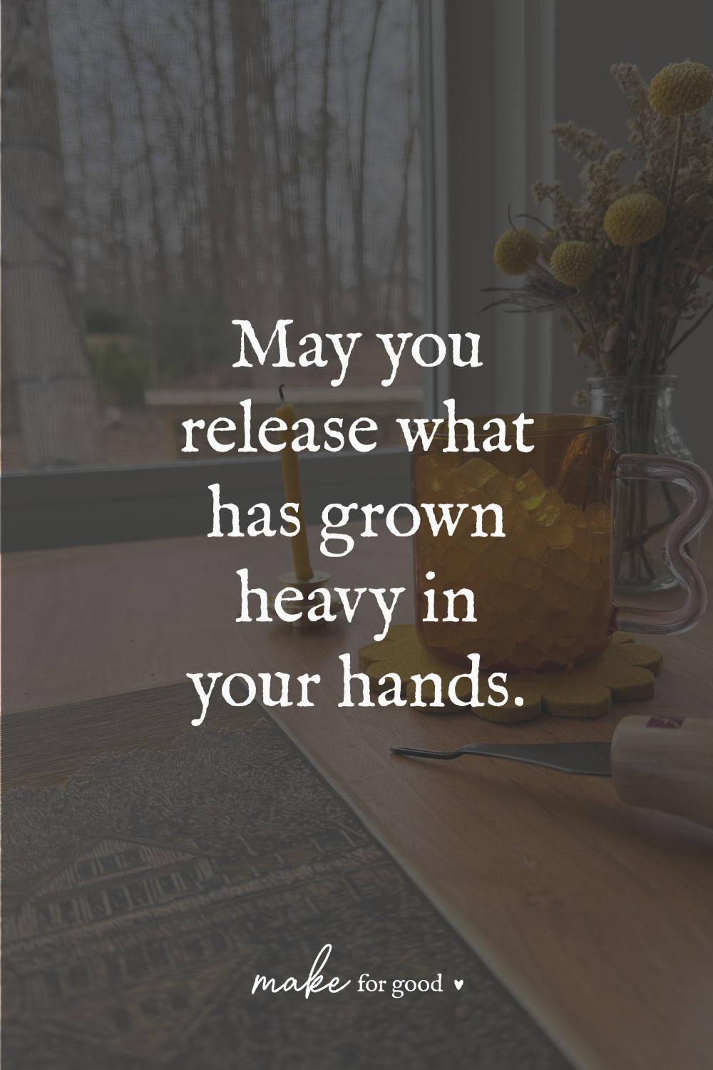 A wooden table by a window with an amber glass mug, a yellow candle, dried flowers, and a linocut carving tool, overlaid with the words “May you release what has grown heavy in your hands.”