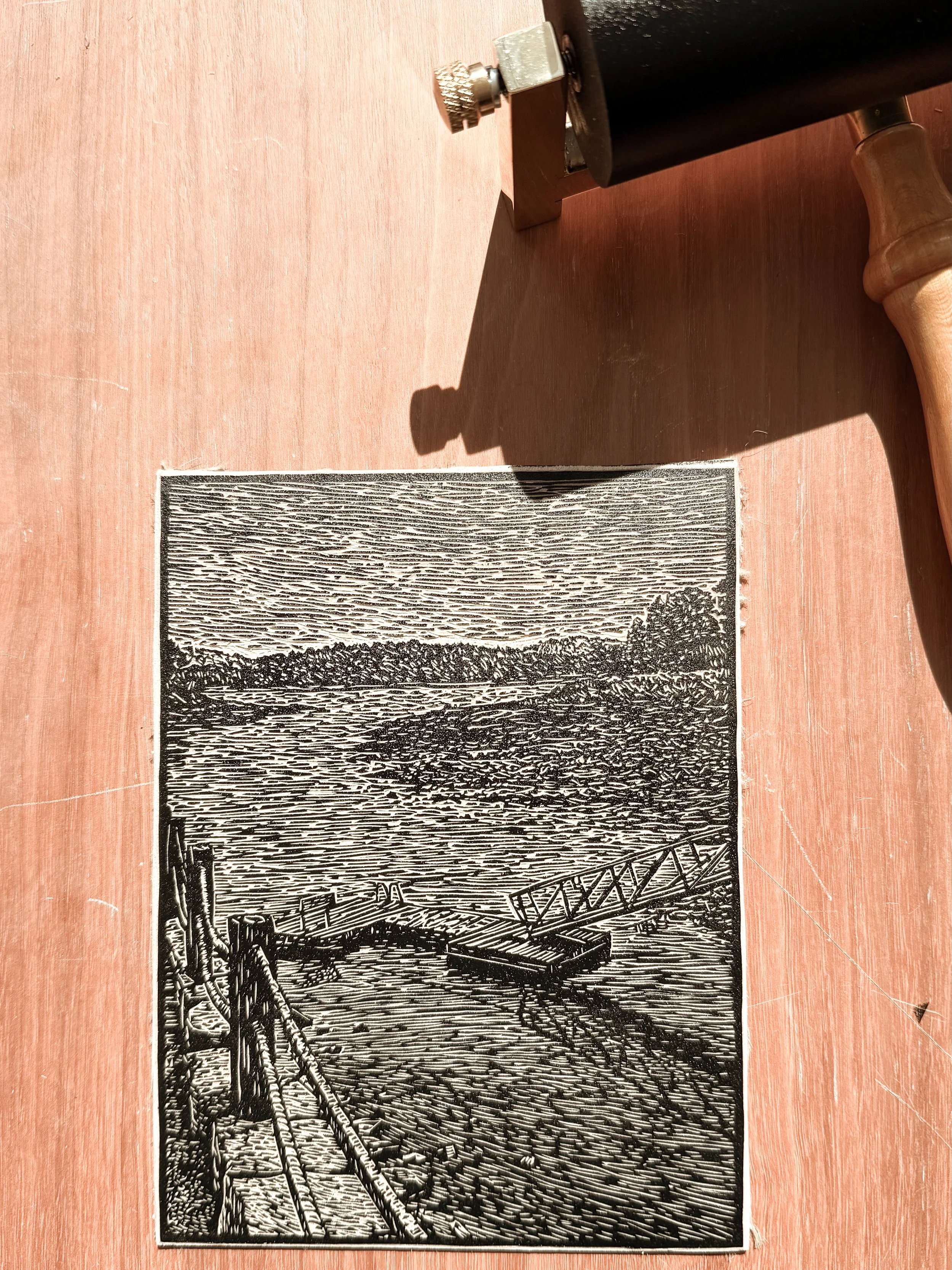 Inked linoleum block with carved coastal dock scene beside brayer, showing printmaking process
