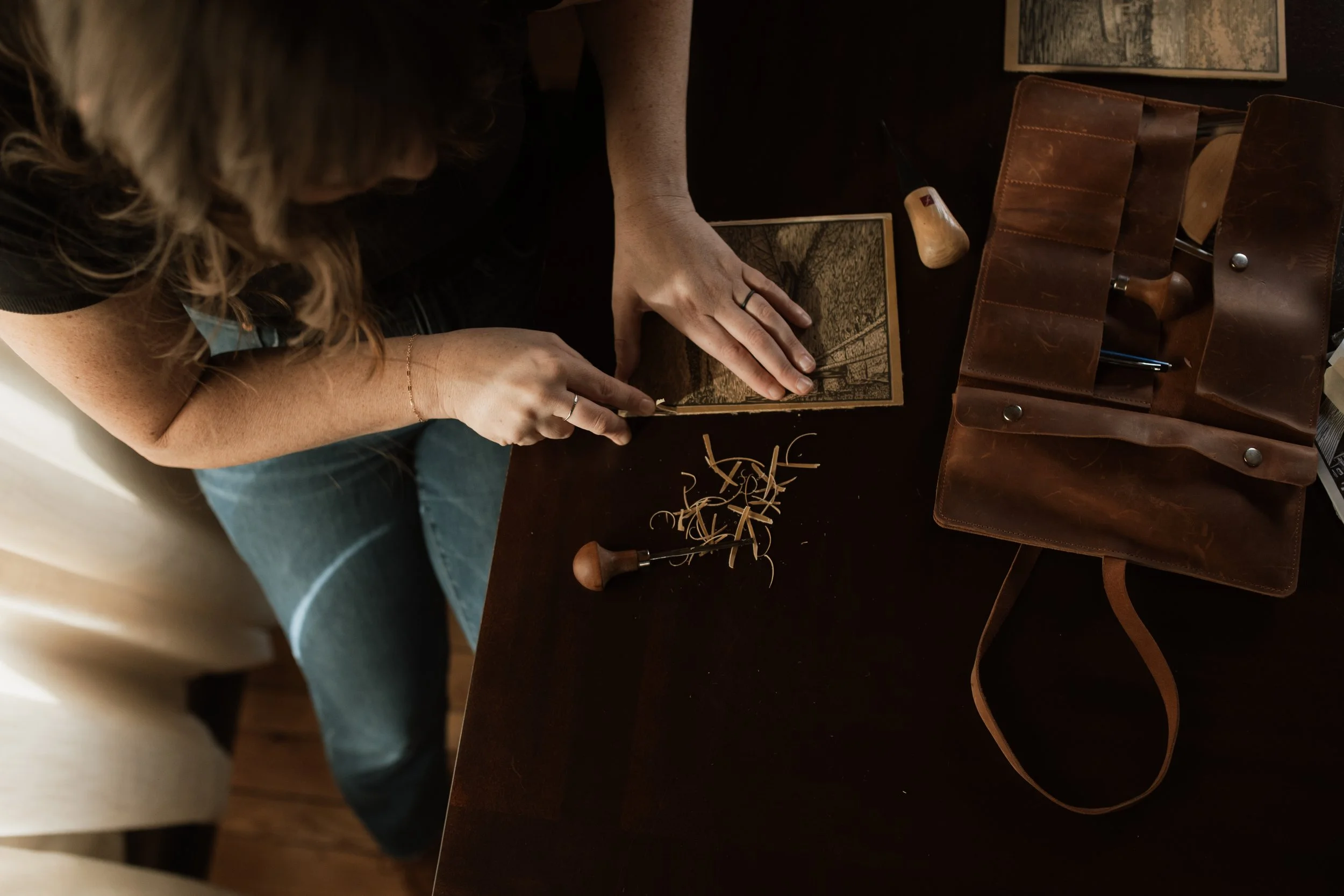 Artist carving a detailed linocut block on a wooden table using fine carving tools