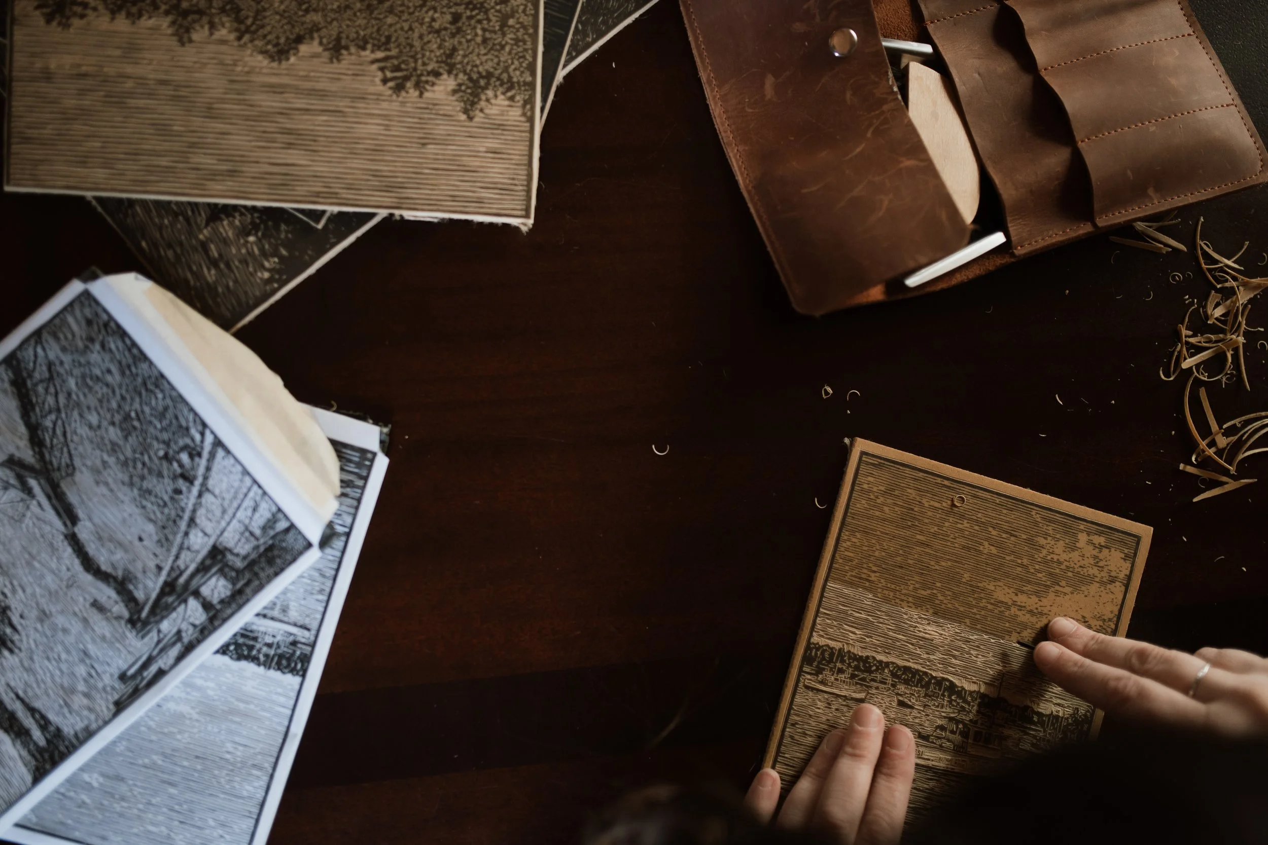 Hands holding a detailed linocut block on a wooden studio table beside a leather tool roll, showing fine carved linework and linoleum shavings from printmaking.
