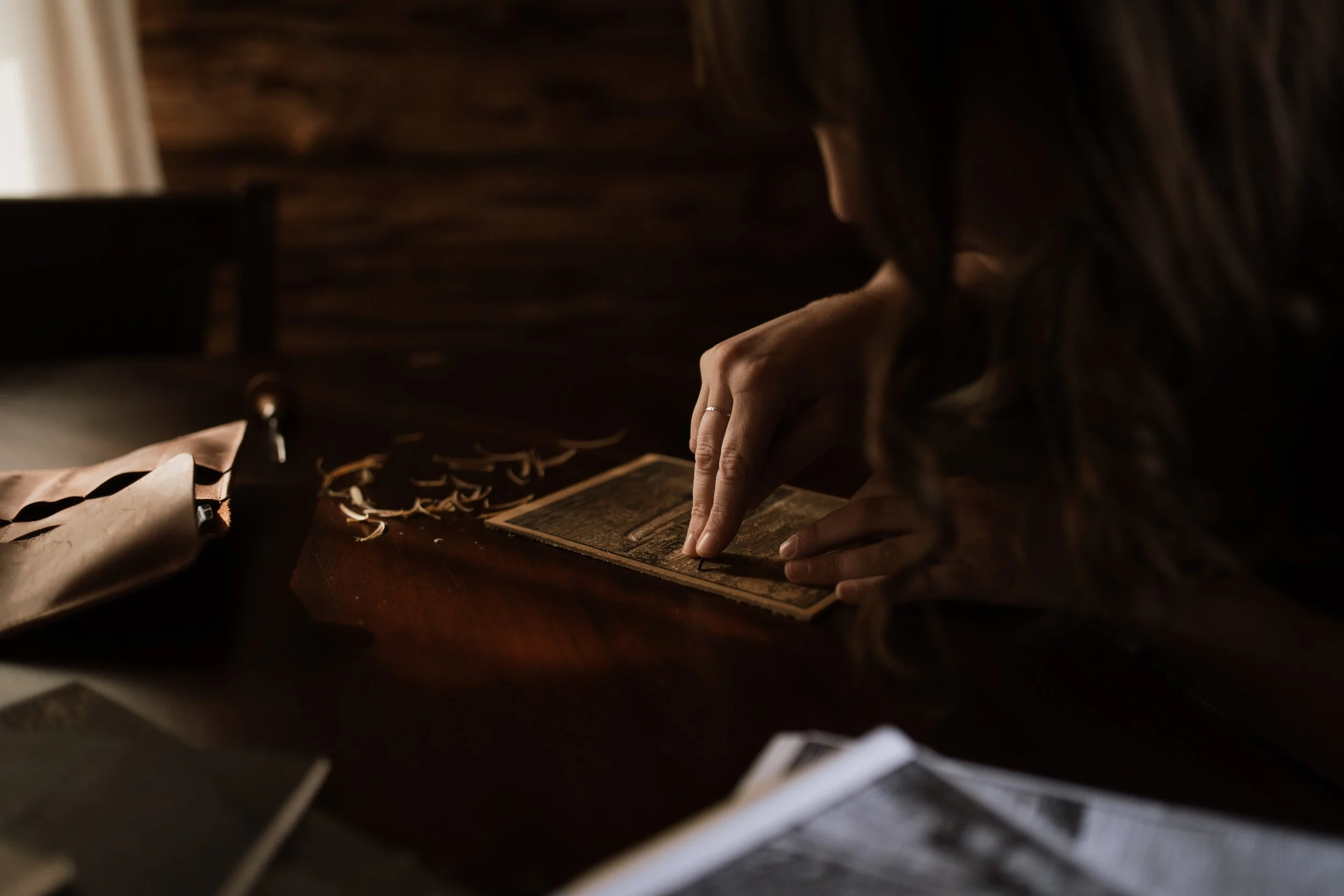 Hand carving a linocut print at a wooden table with carving tools and wood shavings in soft natural light