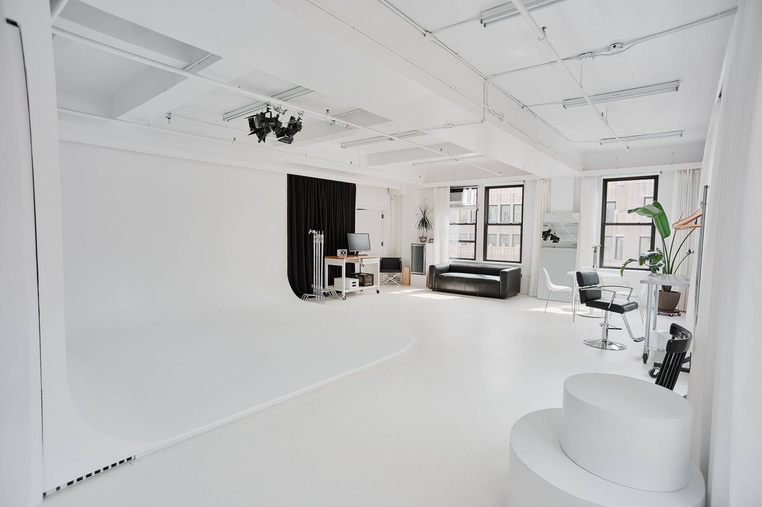 Spacious modern photo studio with white walls, a cyclorama, and lighting equipment. The studio includes a black couch, chairs, desk with computer, potted plants, and large windows.