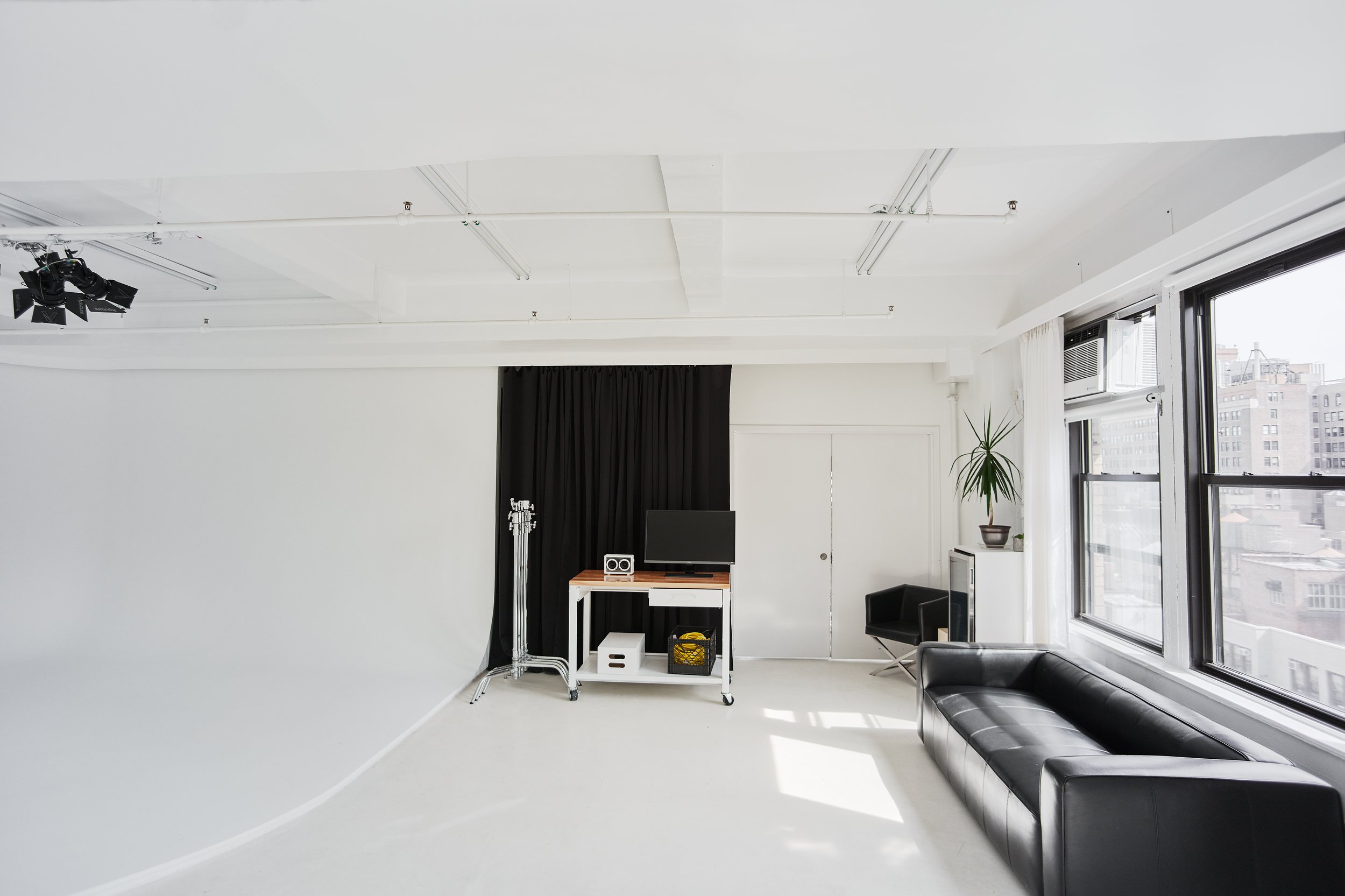 Minimalist photography studio with white walls and a curved white backdrop, black sofa, windows with city view, and lighting equipment.