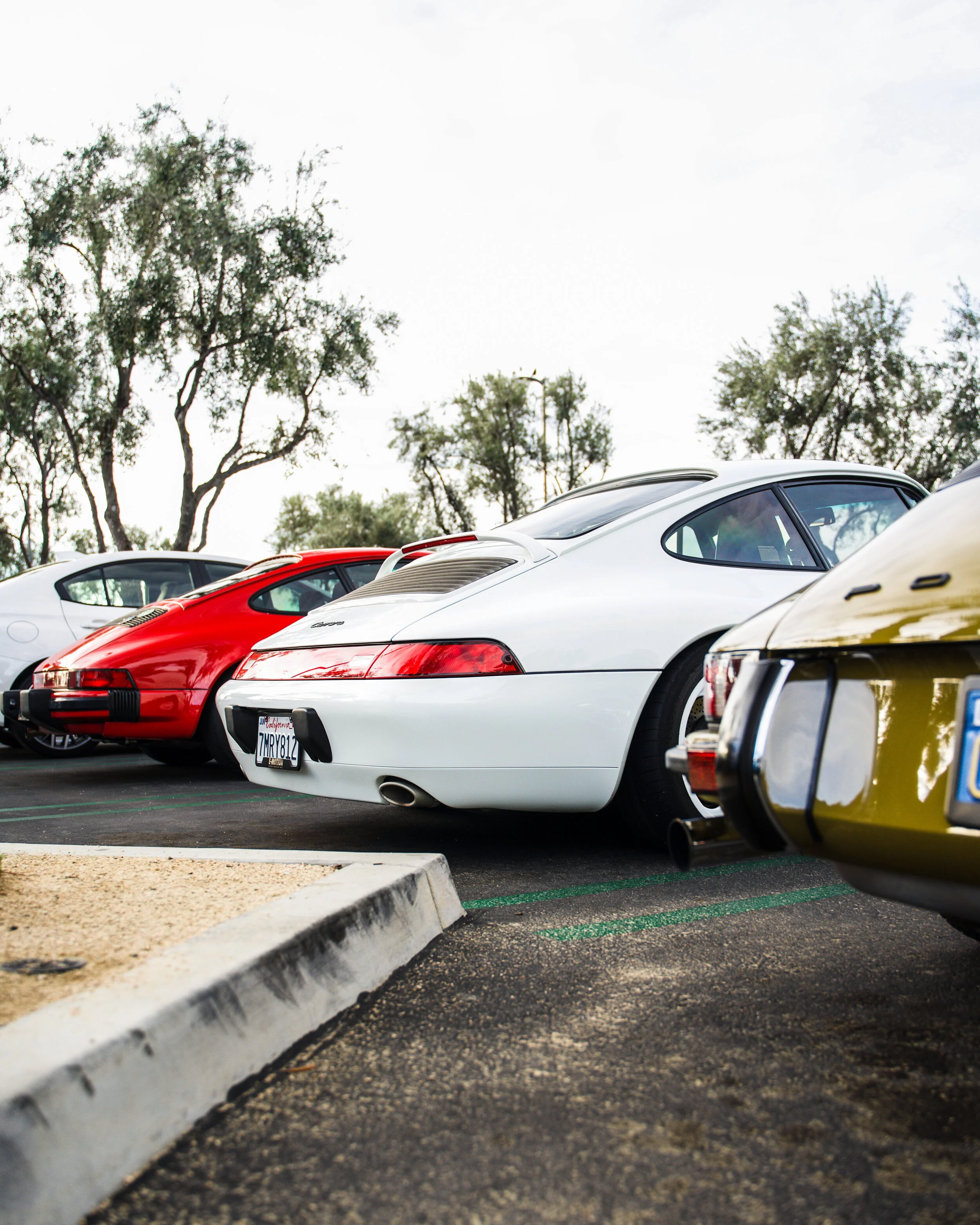 A row of vintage sports cars, including red and white models, parked in a lot with trees in the background.
