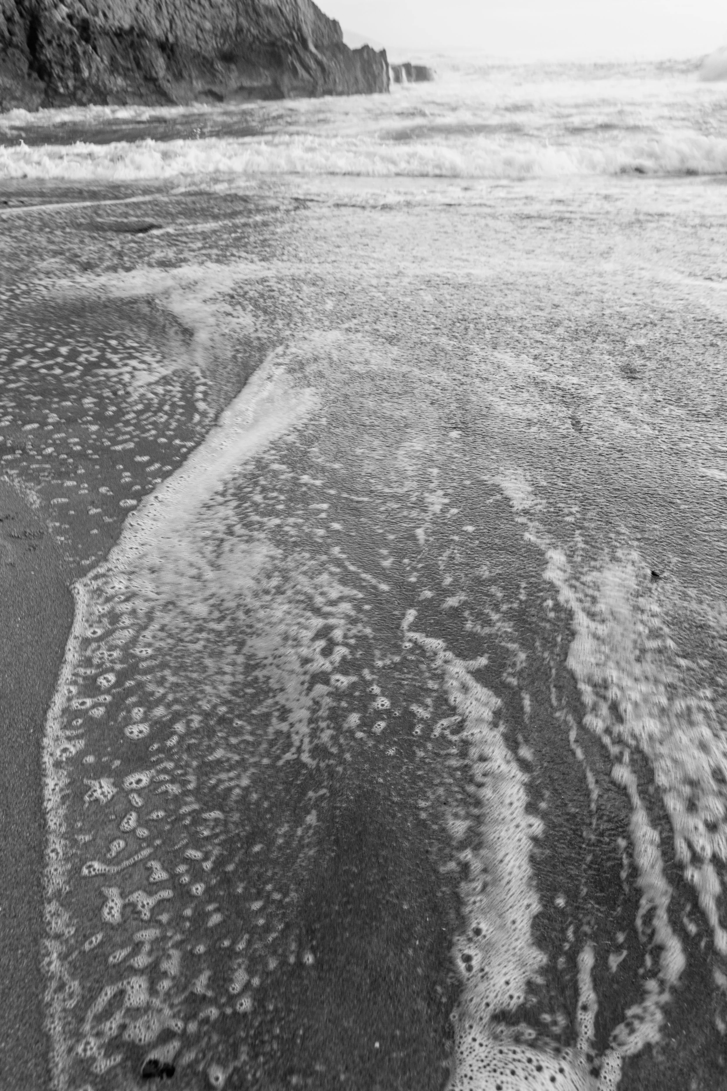 Black and white photo of ocean waves washing up on a sandy beach with rocks in the background.