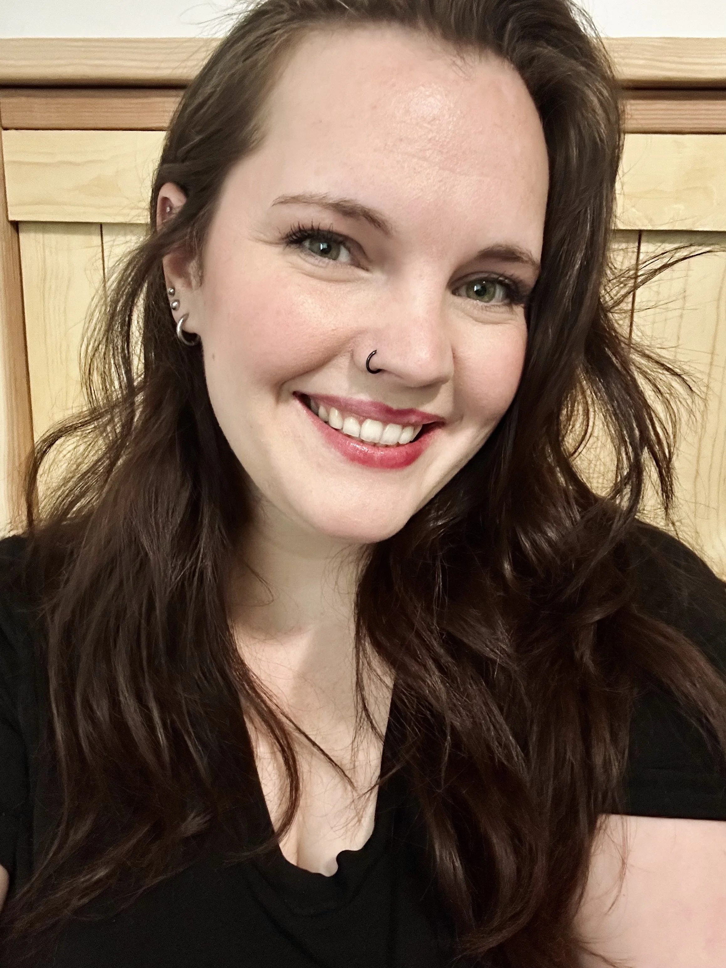 A woman with long brown hair, green eyes, and piercings, smiling at the camera in front of a wooden background.