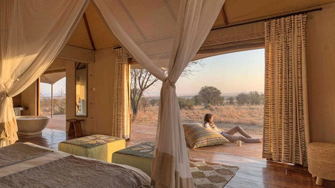 A woman relaxing outside a safari lodge room with open sliding doors, sitting on the wooden deck with a view of the African savannah at sunset.
