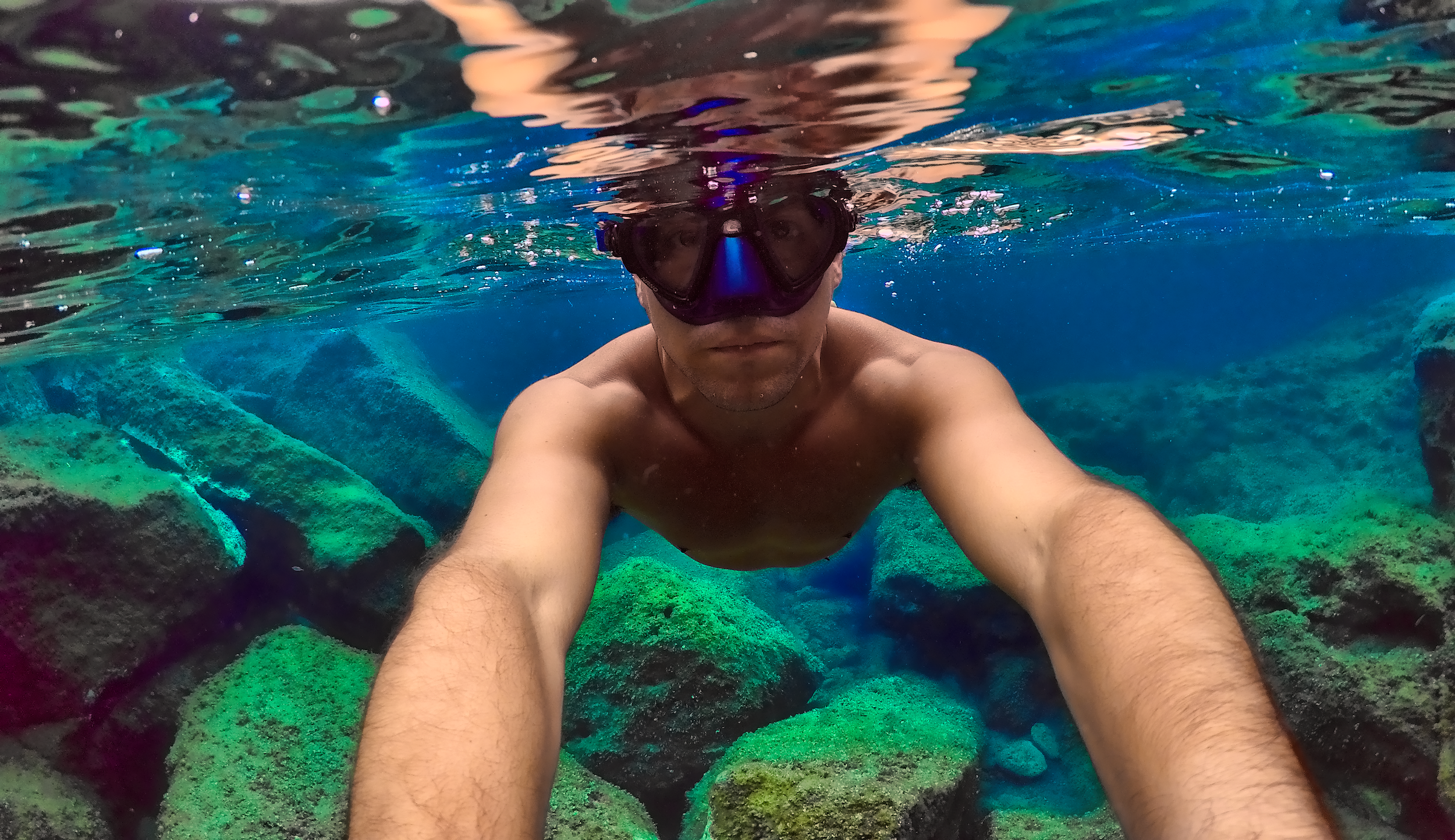 Man taking a selfie underwater, wearing a mask, snorkeling gear, and no shirt, surrounded by rocks and clear blue water.
