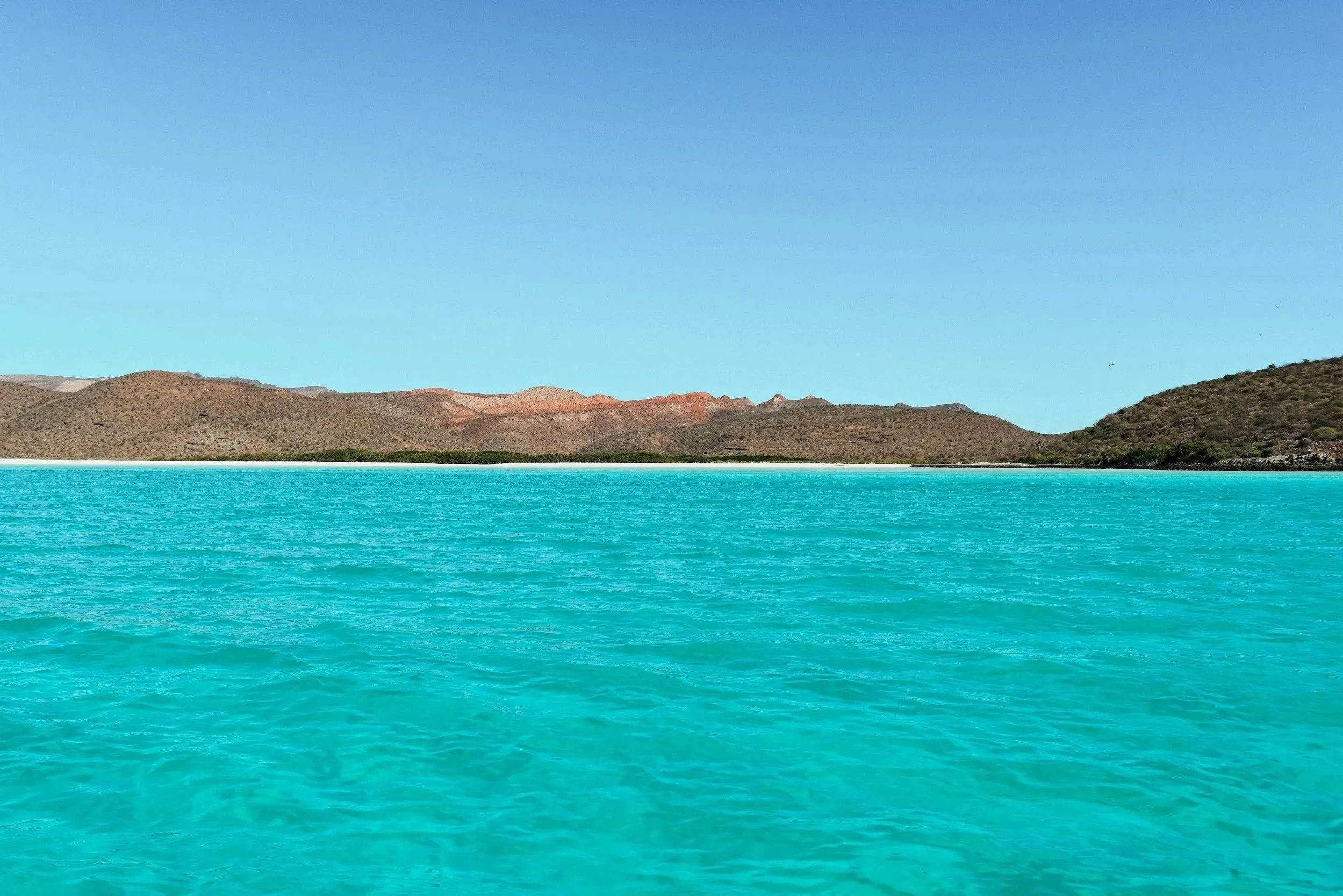 A landscape view of turquoise water, rugged hills, and a clear blue sky.