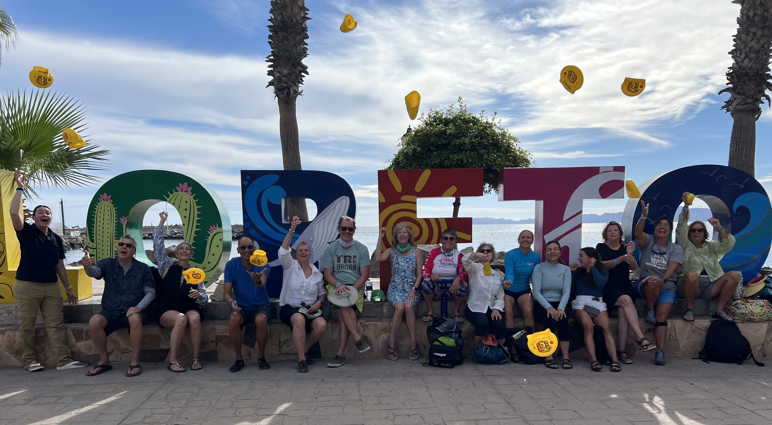 Group of people on the beach sitting and standing in front of large colorful letters spelling 'OTE' with palm trees and clouds in the sky behind them and sea in the background. Some are holding yellow hats, and many are smiling and waving.