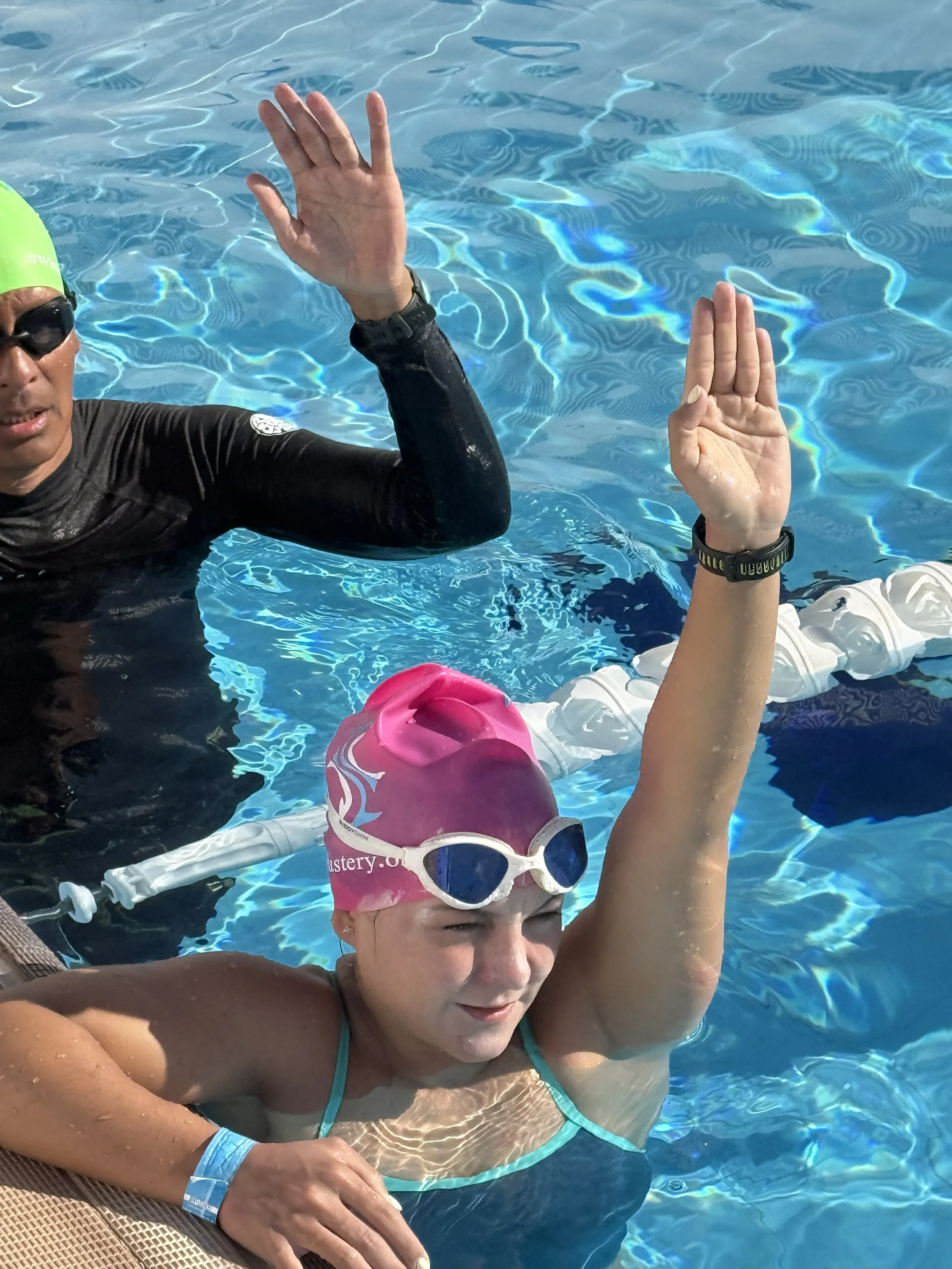 A woman in a swimming pool wearing a pink swim cap and goggles, with a woman in a black wetsuit and a yellow swim cap beside her, both raising their hands for a high five.