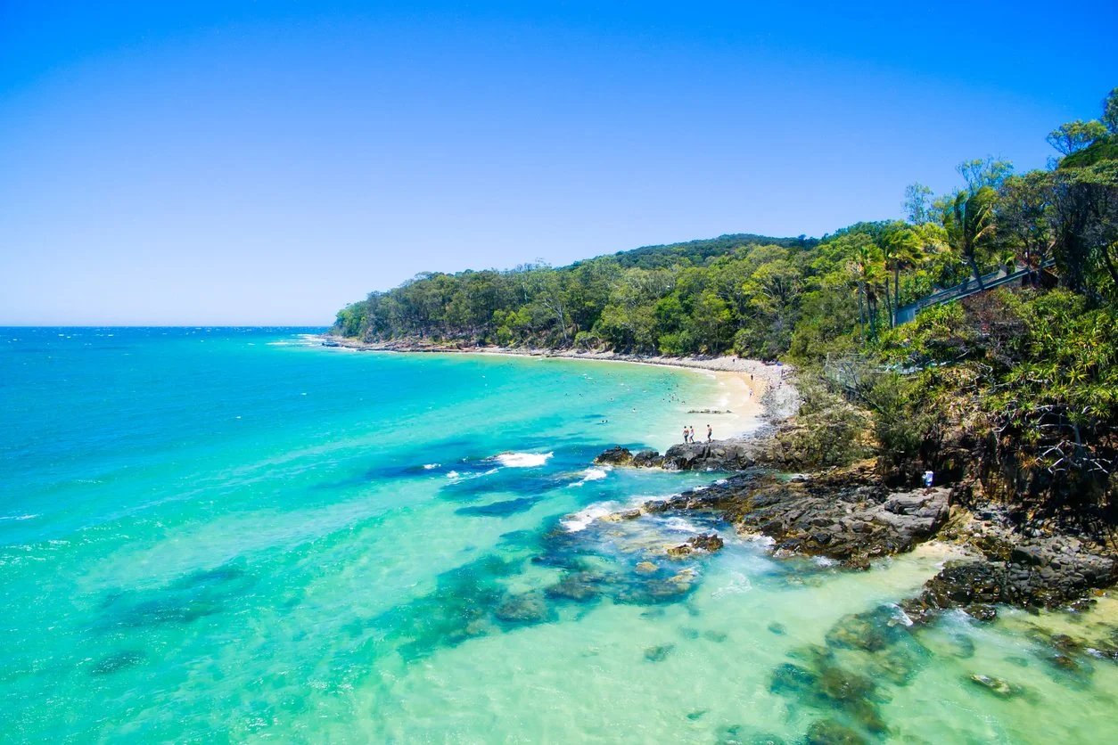 Tropical beach with turquoise water, rocky shoreline, green trees, and a clear blue sky.