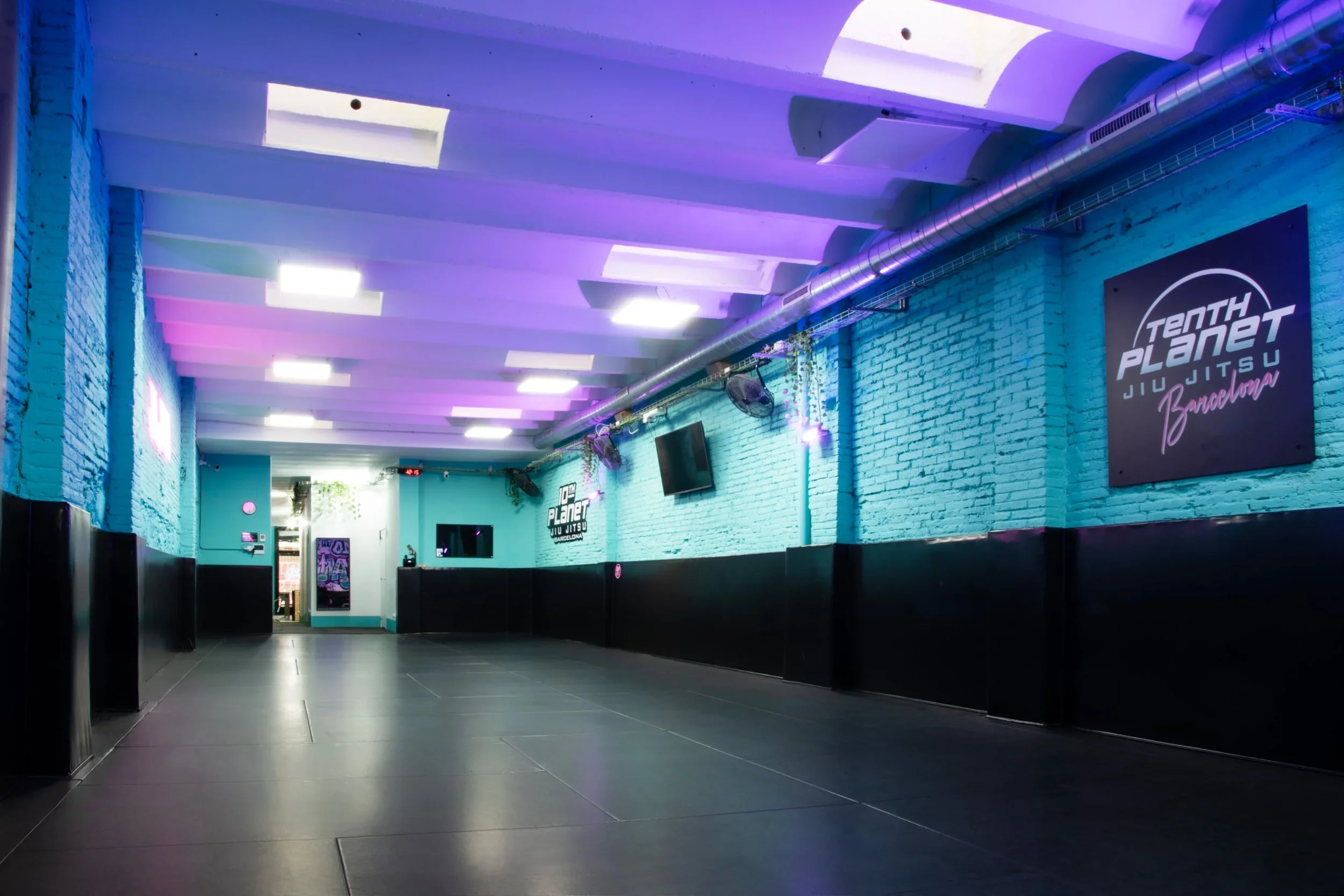 Empty martial arts training room with black mats, teal brick walls, purple ceiling lights, TV screens, and a sign that reads 'Tenth Planet Jiu Jitsu Barcelona'.