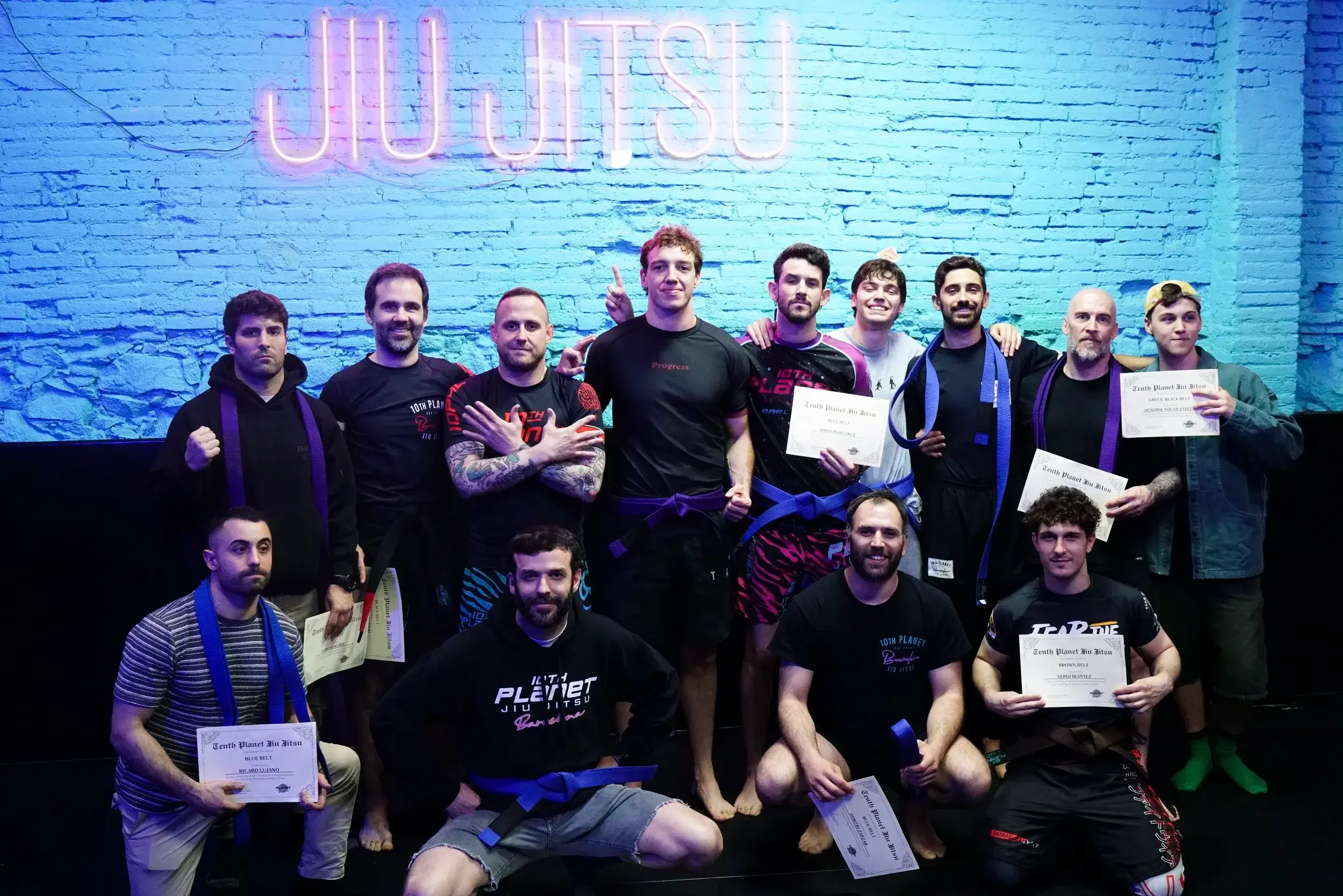 Group of people at a Brazilian Jiu-Jitsu competition, holding certificates and wearing martial arts uniforms with belts, standing in front of a brick wall with a neon sign reading 'Jiu Jitsu'.