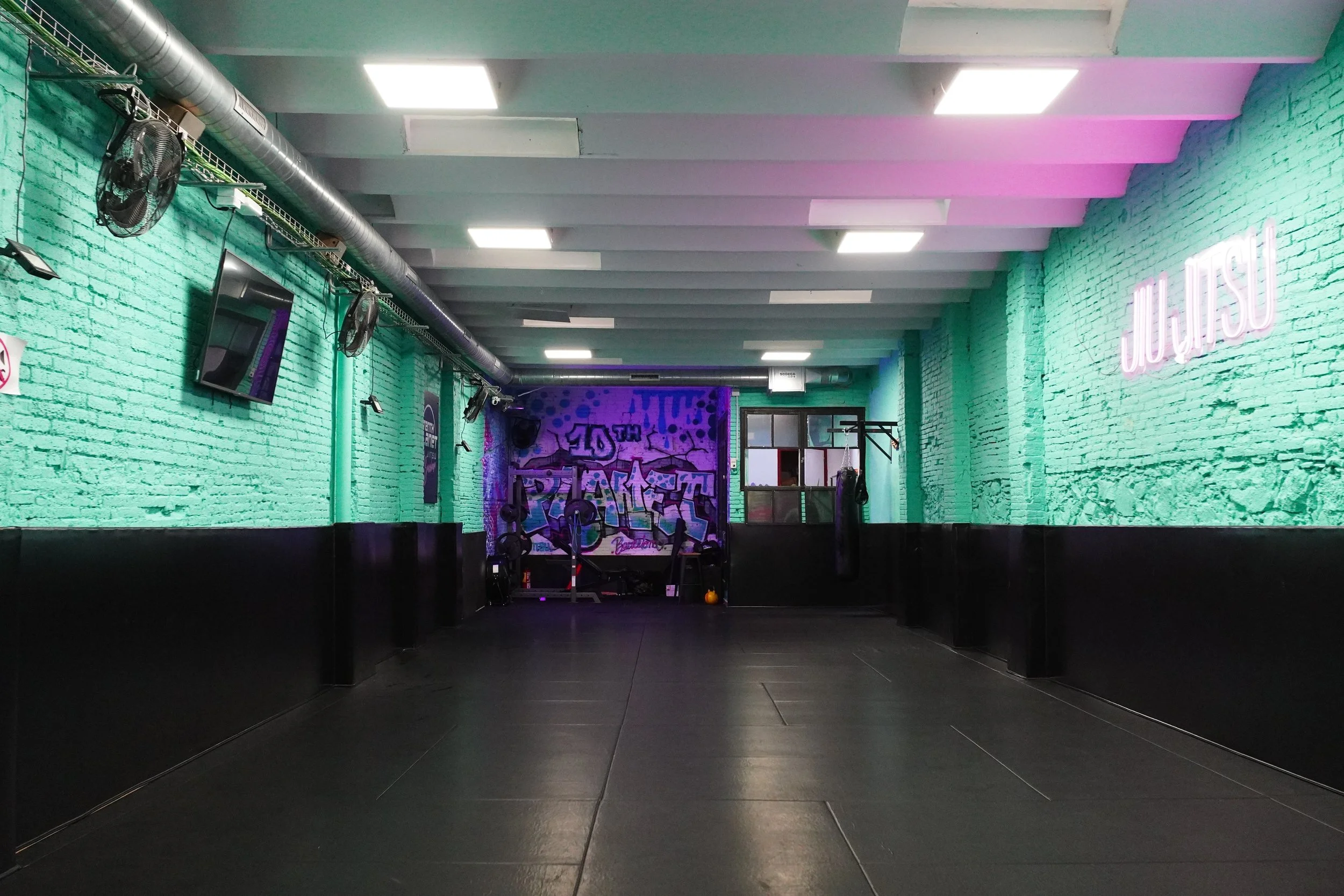An empty gym with green brick walls, black flooring, and neon signs. Punching bags and fitness equipment are visible at the back, with graffiti art on a purple wall in the background.