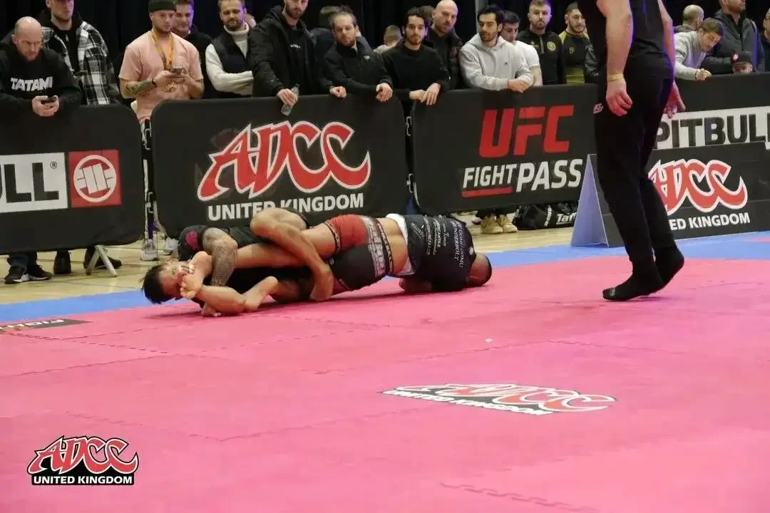 Two mixed martial arts fighters grappling on the mat during a match, with a referee observing nearby, and spectators watching behind a barrier.