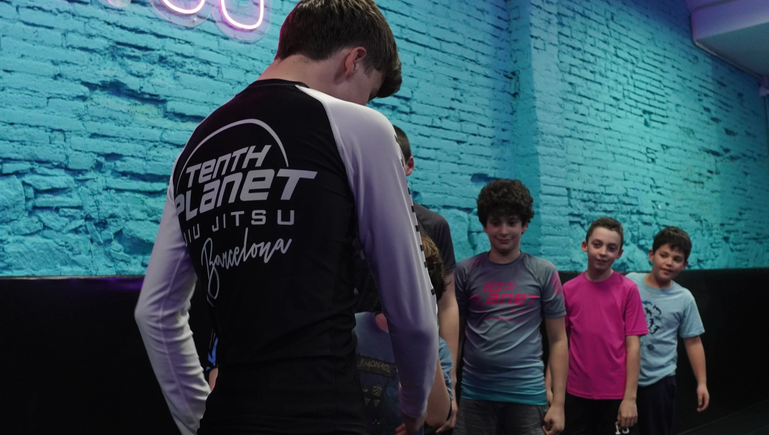 A group of children standing indoors against a turquoise brick wall with neon lighting, with a teenage boy in the foreground wearing a black and white shirt that says "Tenth Planet Jiu Jitsu Barcelona." The children are smiling and looking at the boy.