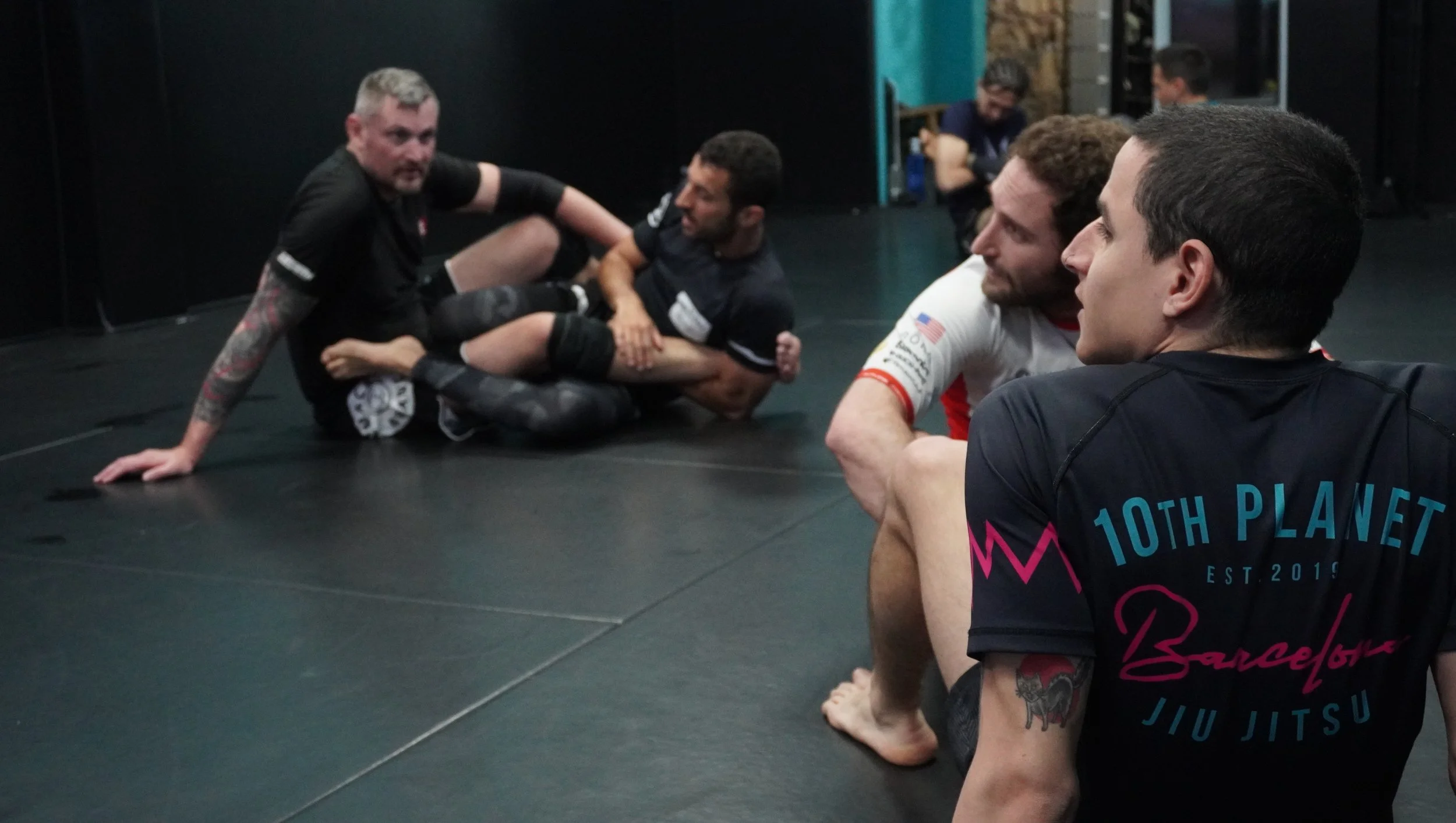 Group of people in a Brazilian Jiu-Jitsu class on the mat, engaging in grappling techniques, with some observing in the background.
