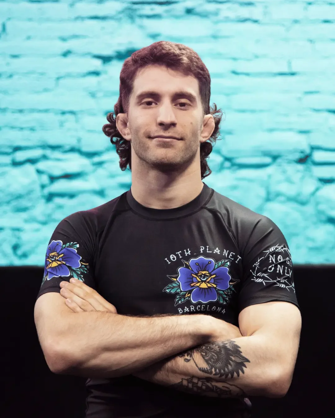 A man with shoulder-length curly brown hair, crossed arms, wearing a black athletic shirt with colorful floral designs and the words "Loth Planet Barcelona" on the front, stands in front of a blue-tinted brick wall backdrop.