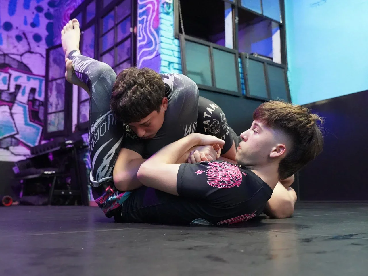 Two men practicing Brazilian Jiu-Jitsu on a mat, with one man in a black gi and the other in a rash guard, in an indoor facility with graffiti-style wall art.