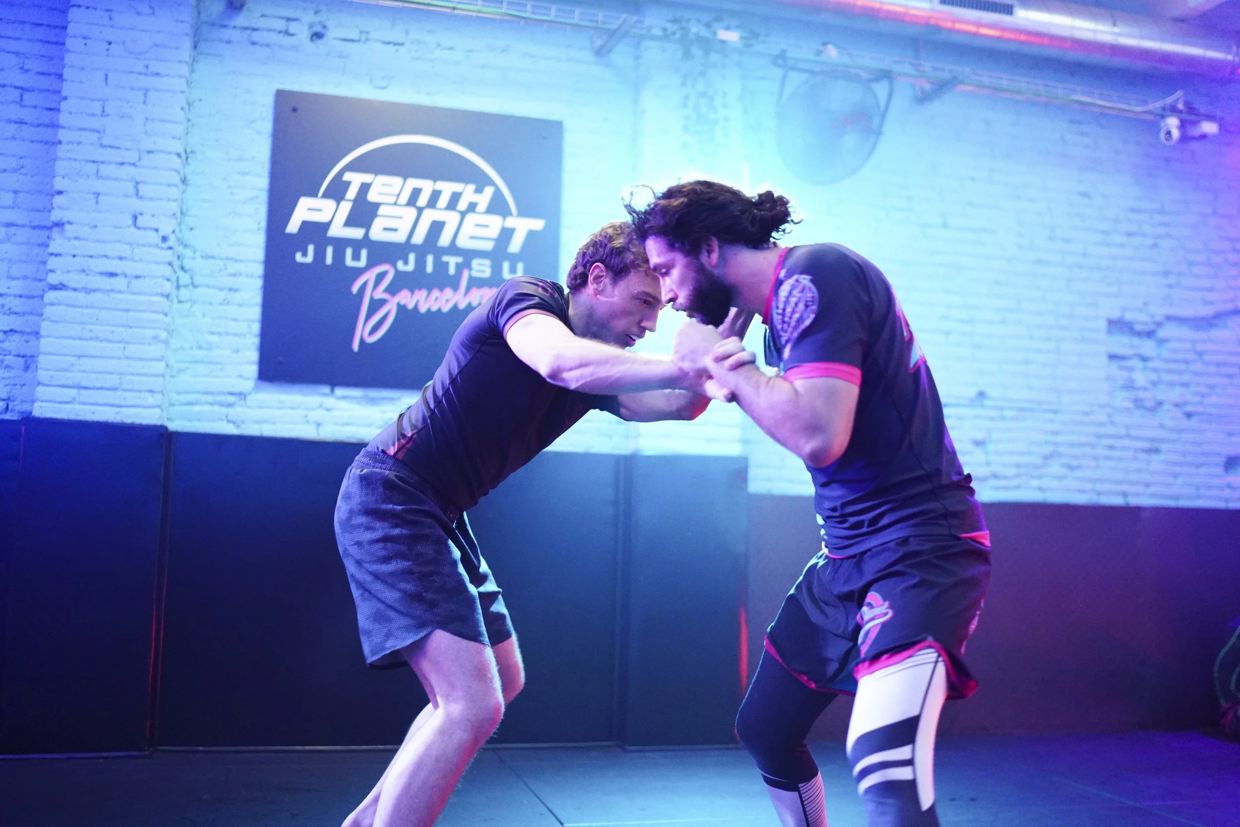 Two men practicing Brazilian Jiu-Jitsu in a gym with a sign that reads 'Tenth Planet Jiu Jitsu Bangkok' on the wall, engaging in a grappling hold.
