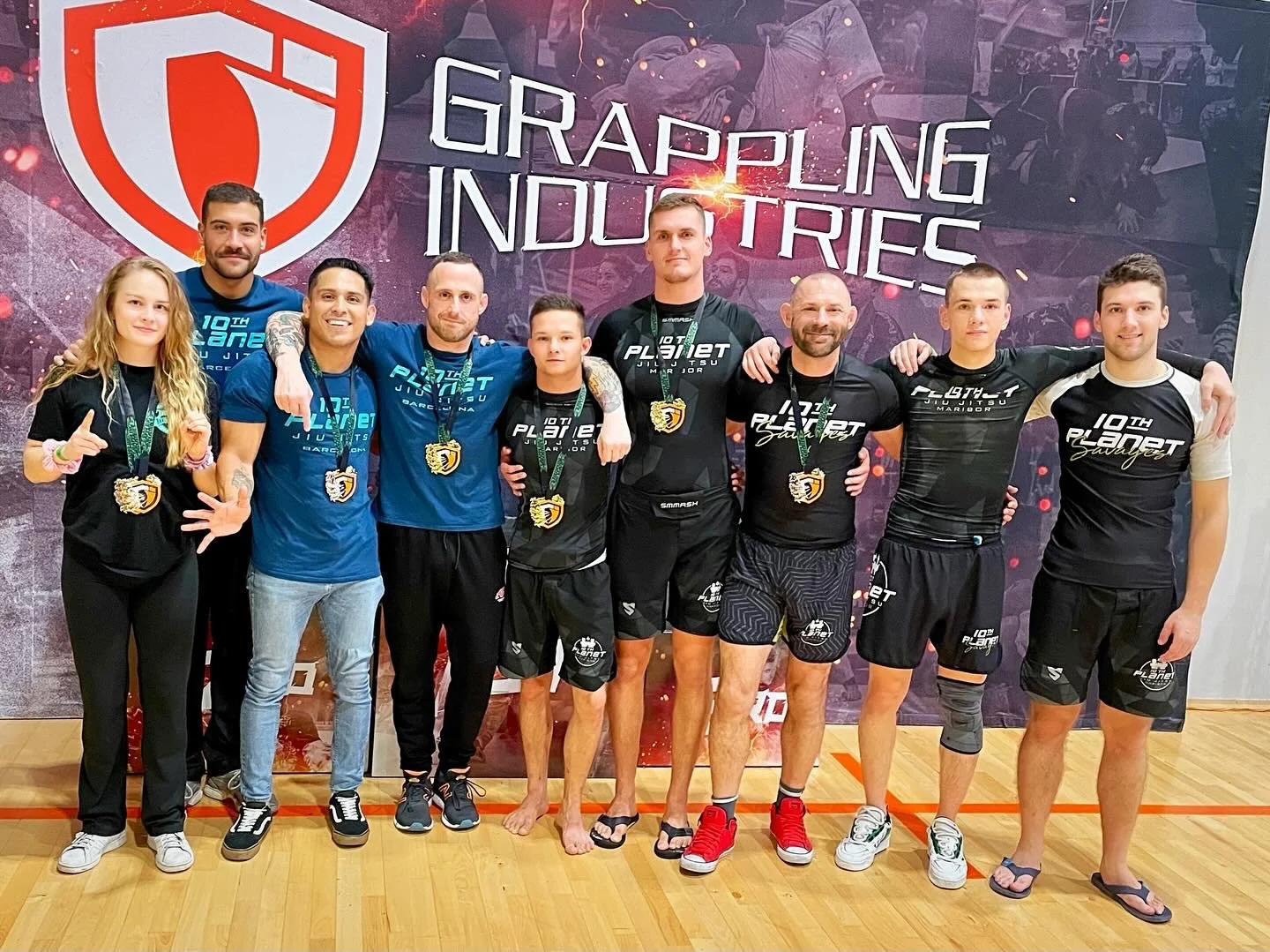 Eight people, seven men and one woman, stand in a gymnasium in front of a large banner that reads 'Grappling Industries'. They are wearing medals around their necks and athletic clothing, celebrating their victory at a grappling or Brazilian Jiu-Jitsu competition.