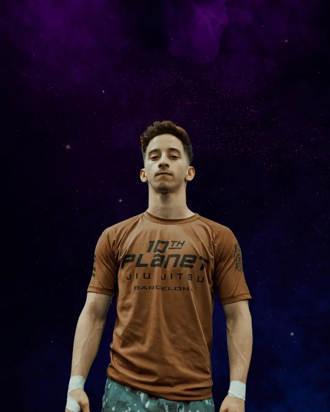 Young man standing against a starry galaxy background, wearing a brown T-shirt with black text and camouflage shorts.