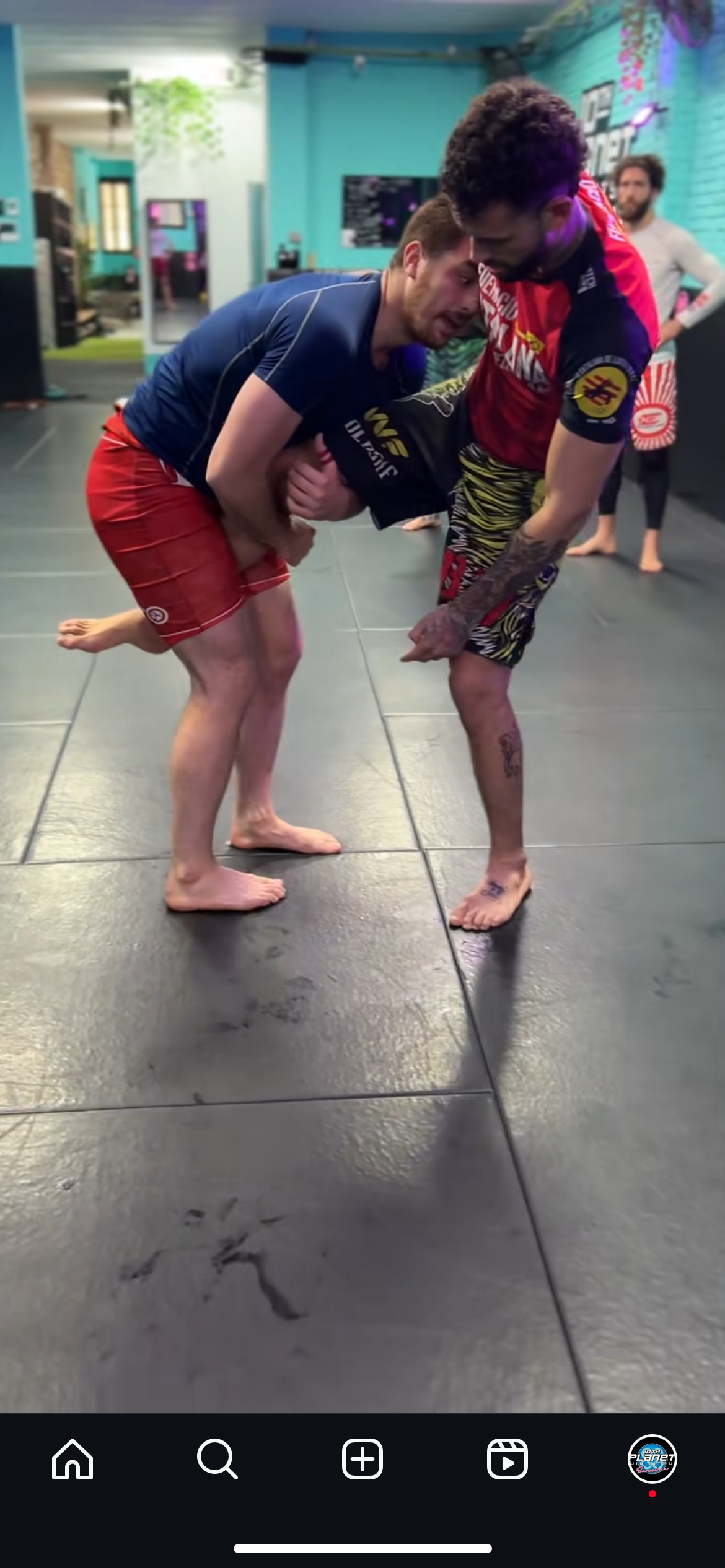 Two men practicing Brazilian Jiu-Jitsu on a mat in a gym, one in a blue shirt and red shorts, the other in a red and black rash guard and multicolored shorts, with a third person watching in the background.