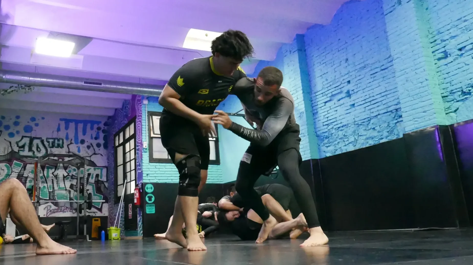 Two men practicing Brazilian Jiu-Jitsu in front of an audience in a gym with blue lighting and a brick wall, with spectators sitting on the floor and standing around.