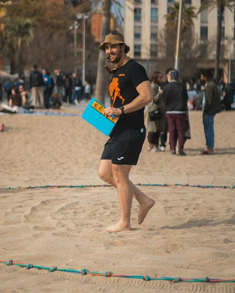 A smiling man wearing a brown hat, sunglasses, a black T-shirt with orange graphic, and black shorts walking barefoot on the sand at the beach, holding a blue and yellow item, with other people and buildings in the background.