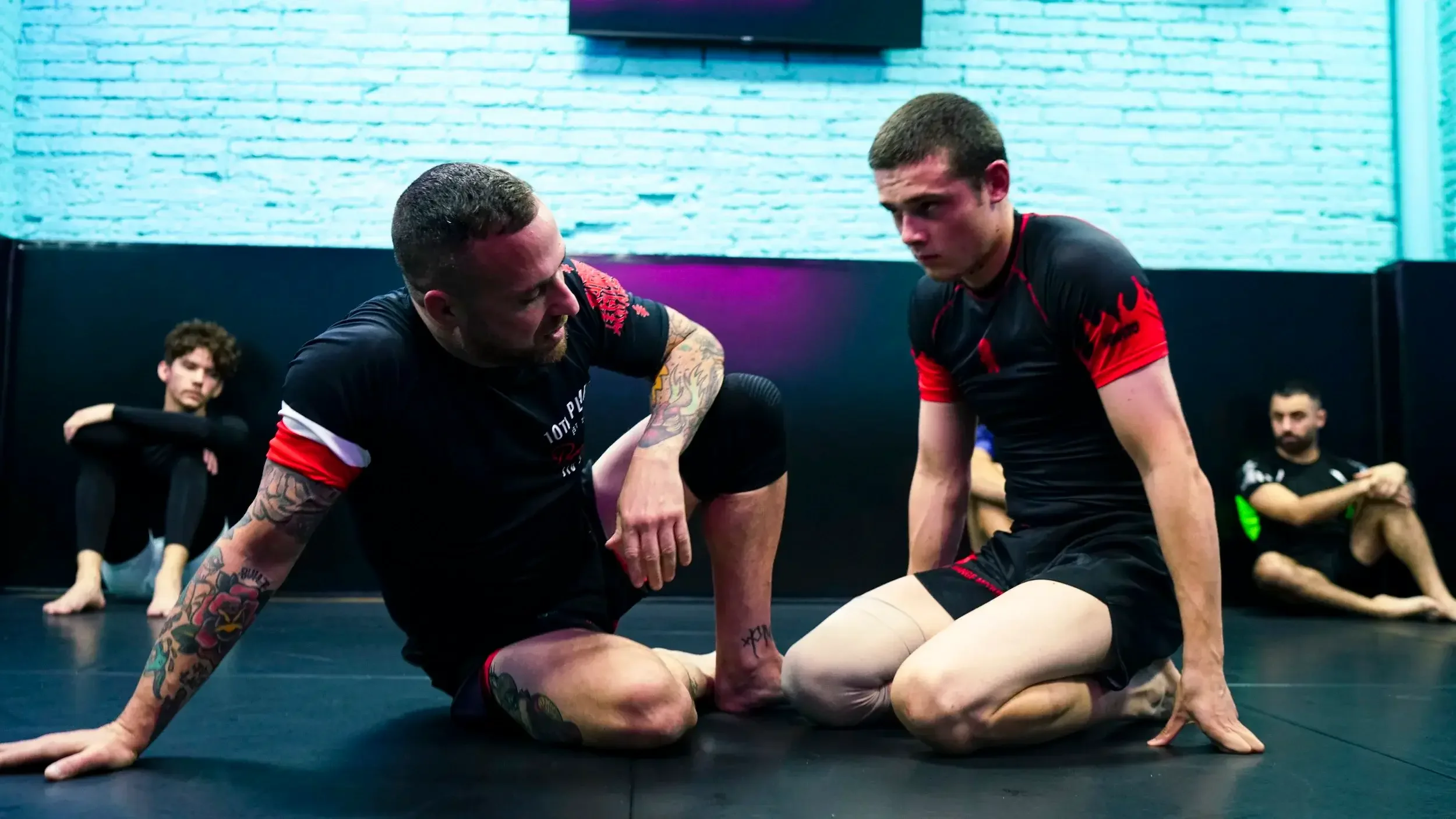 Two men dressed in martial arts shorts and shirts are kneeling on the mat with one leaning forward with his arm wrapped around the other's neck, practicing Brazilian Jiu-Jitsu techniques. Two other individuals are seated in the background against a black wall, observing or resting.