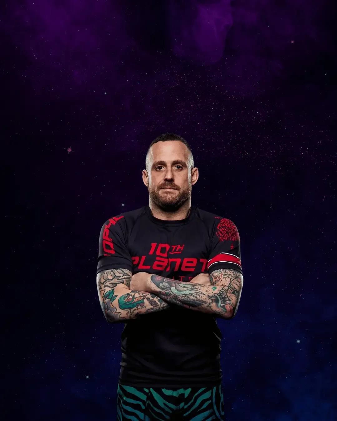 A man with tattoos on his arms and wearing a black T-shirt with red text standing with his arms crossed in front of a starry space background.